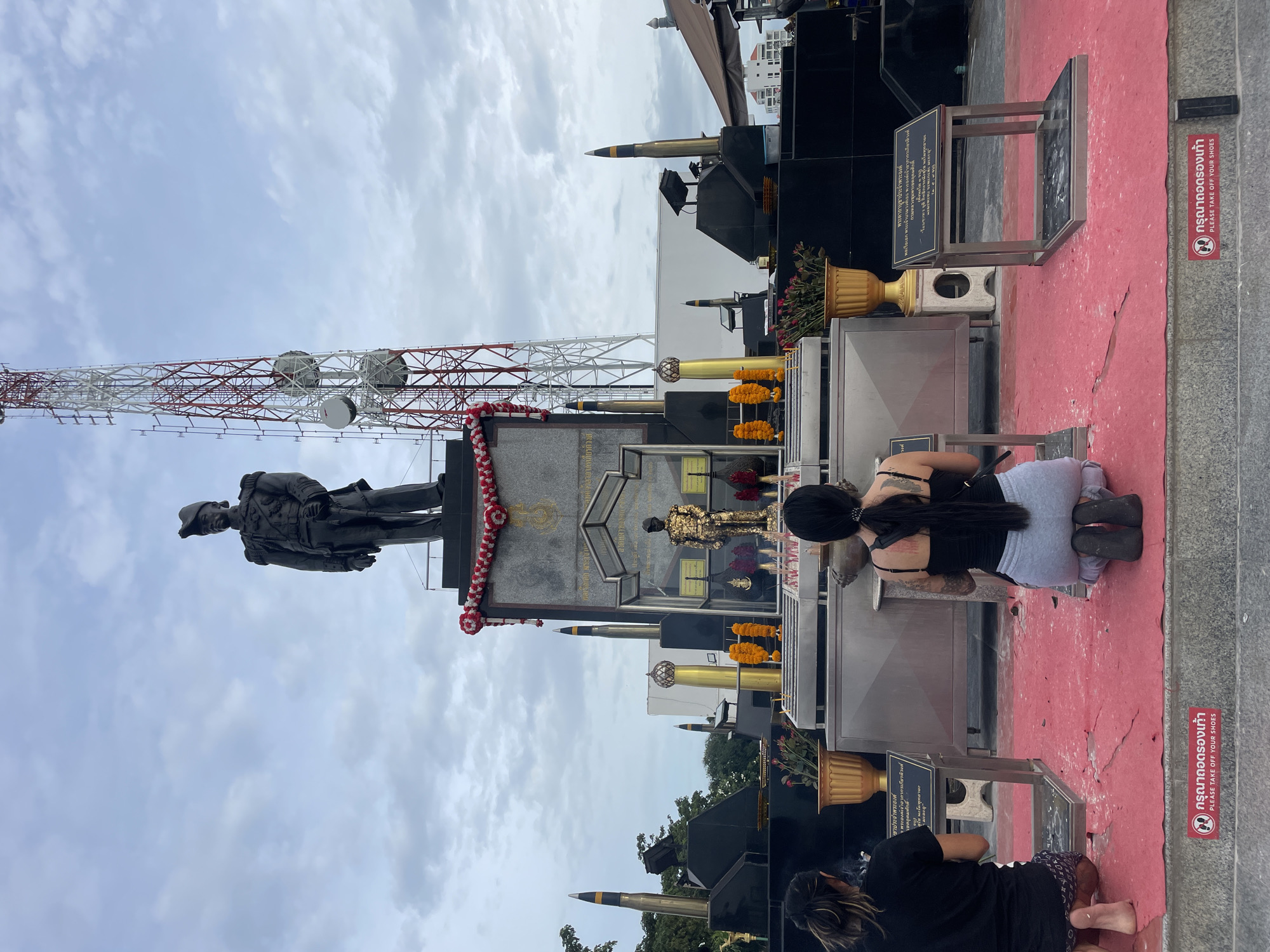 Two individuals are kneeling on a red carpet in front of a monument in Pattaya City, Thailand. The monument features a large statue of a man in uniform at the top, with a smaller golden statue in front of a dark grey base. Surrounding the base are two large missile-like objects, a tall radio tower, and various decorative elements including garlands and potted plants. The individuals are facing the monument, with one person wearing a black t-shirt and the other a black tank top and grey sweatpants. Both are engaged in prayer or meditation, with incense burning. A sign at the bottom of the image instructs visitors to "PLEASE TAKE OFF YOUR SHOES." The sky is overcast.