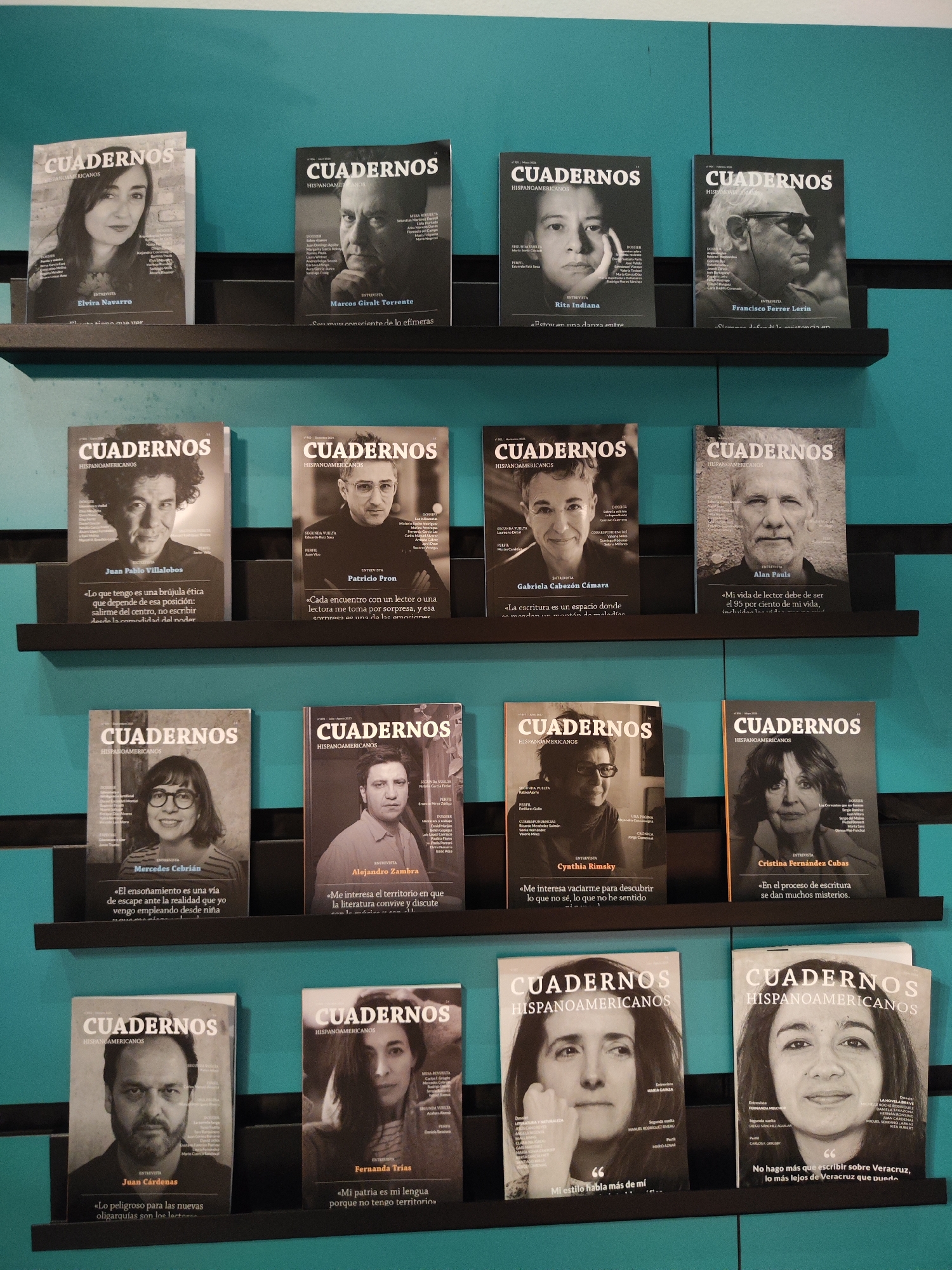 This image depicts sixteen copies of "Cuadernos Hispanoamericanos" magazines displayed on a teal wall in Providencia, Chile. The magazines are arranged in four horizontal rows of four, resting on slim black shelves. Each magazine cover features a black and white portrait of a different individual, with the title "CUADERNOS HISPANOSAMERICANOS" prominently displayed in white text at the top. Below the title, "ENTREVISTA" (Interview) is visible, followed by the name of the featured individual. Some covers also include a short quote attributed to the interviewee.
From top to bottom, left to right, the identifiable individuals on the covers are:
* **Row 1:** Elvira Navarro, Marcos Giralt Torrente, Rita Indiana, Francisco Ferrer Lerín.
* **Row 2:** Juan Pablo Villalobos, Patricio Pron, Gabriela Cabezón Cámara, Alan Pauls.
* **Row 3:** Mercedes Cebrián, Alejandro Zambra, Cynthia Rimsky, Cristina Fernández Cubas.
* **Row 4:** Juan Cárdenas, Fernanda Trias, María Gainza, Fernanda Melchor.
The wall behind the display is a solid, uniform teal color. The shelves are black and extend horizontally across the frame. The lighting is even, illuminating the magazines clearly without harsh shadows. No individuals are physically present in the scene, only their portraits on the magazine covers. The overall presentation suggests a curated literary display within an indoor environment.
