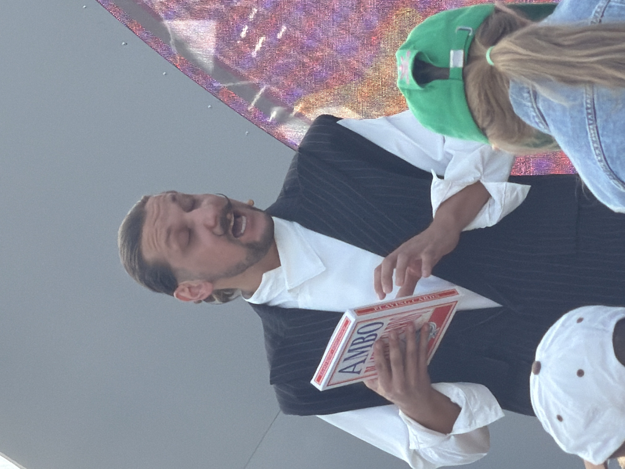 An outdoor performance scene captured in Kazan, Russia, features a male performer addressing an unseen audience. The man, positioned centrally, has dark hair slicked back, a prominent handlebar mustache, and a short beard. He wears a white long-sleeved shirt with a high, structured collar, topped by a dark, pinstriped sleeveless vest or jacket. His mouth is open as if speaking or singing, and his eyes are narrowed. In his left hand, he holds a red and white box, likely a prop or deck of playing cards, with the word "AMBO" clearly visible in red letters on its white face. His right hand is raised slightly, gesturing.
In the lower right foreground, the back of an audience member's head and upper torso are visible. This individual has light brown hair tied in a ponytail with a light green hair tie and wears a green baseball cap and a denim jacket. In the lower left foreground, only the crown of another audience member's head is visible, wearing a white baseball cap with small brown details.
The background on the left consists of a plain, light grey or blue wall or sky. To the right, a large, pixelated digital screen or display panel shows a vibrant, abstract pattern in hues of purple, pink, and white. Several small, reflective dots are scattered across its surface.
