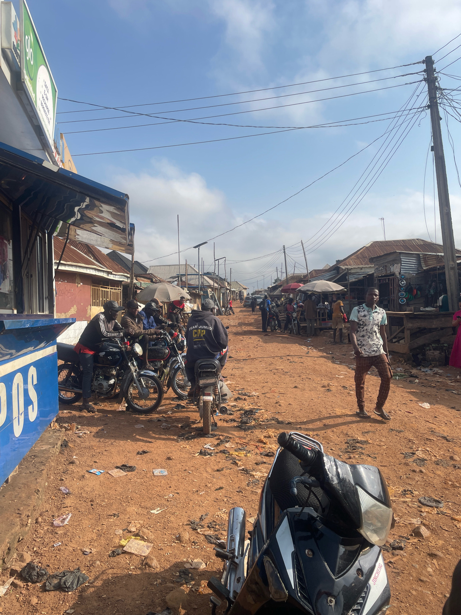 The image depicts a street scene in Kurunduma, Nigeria. The street is unpaved and dusty, with a reddish-brown dirt surface. On the left side of the image, there is a building with a blue and white facade, which appears to be a commercial establishment. Several motorcycles are parked in front of this building, with a group of men gathered around them. The men are dressed in casual clothing, and some are wearing caps.
The street is lined with small shops and stalls on both sides, many of which have corrugated metal roofs. A utility pole with numerous wires runs along the right side of the street. Several people are walking or standing around, engaged in various activities. One man in a patterned shirt and checkered pants is walking towards the camera in the foreground. Another man in a black hooded sweatshirt is standing near a motorcycle. Umbrellas are set up over some of the stalls, providing shade.
The overall impression is of a busy, informal market or commercial area in a Nigerian town. The sky is partly cloudy, with a clear blue sky visible in some sections. The ground is littered with some debris, including plastic bags and wrappers.
