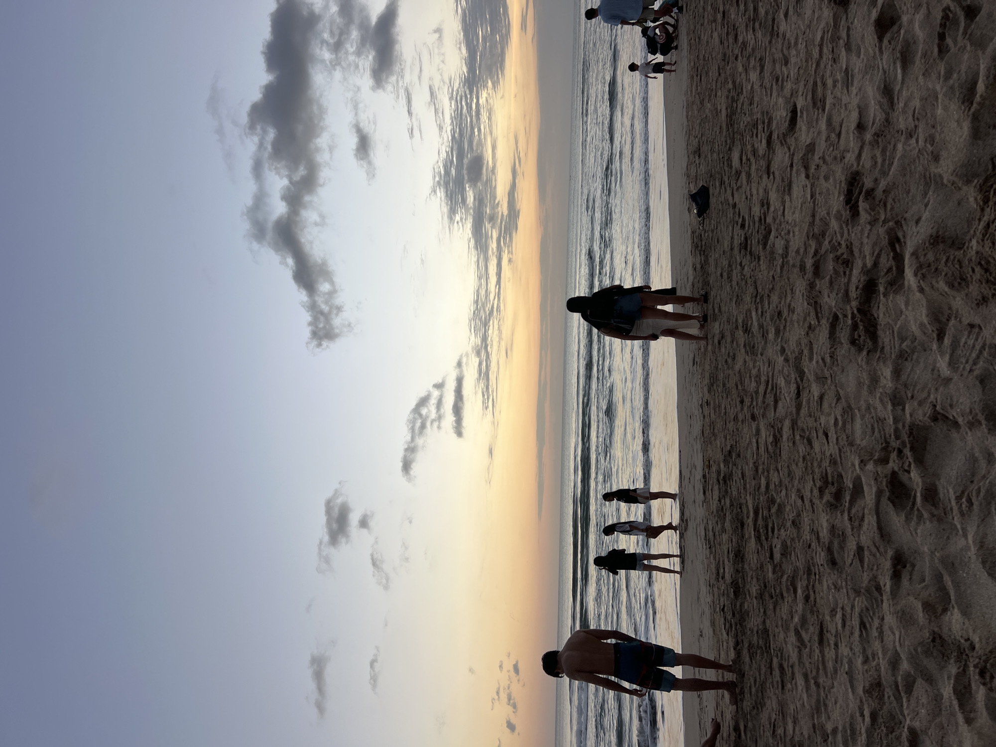 At dusk on Kuta Beach in Indonesia, a diverse group of individuals enjoys the shoreline. The sky, a gradient of soft blues and warm oranges, is dotted with scattered clouds. The ocean, calm with gentle waves, meets the sandy beach, where footprints mark the wet sand.
In the foreground, a lone figure, silhouetted against the horizon, stands with his back to the camera, wearing dark shorts. Further down the beach, several other figures are visible. Three individuals, appearing to be children or young adults, stand near the water's edge, looking out at the ocean. Closer to the foreground, two adults stand together, one with their arm around the other, facing the sea.
To the right, a young child walks towards the water, accompanied by two adults. One adult, wearing a light blue shirt, stands nearby, while another is crouched down, seemingly attending to something on the ground. A dark bag lies on the sand between the two groups of people. The overall scene evokes a peaceful evening at a popular tourist destination.