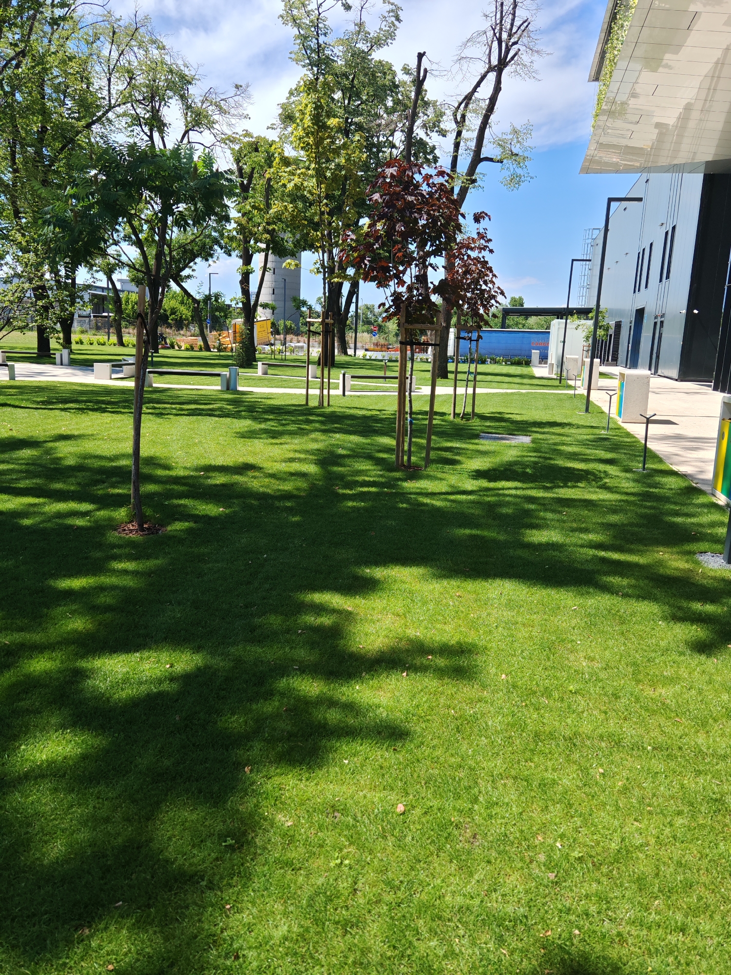 The image depicts an outdoor scene in a modern urban park setting, likely in Plovdiv, Bulgaria, adjacent to a contemporary building. The foreground is dominated by a lush, well-maintained green lawn, with dappled sunlight casting shadows of trees across its surface. Several young trees, some with dark red foliage, are planted at intervals, supported by wooden stakes.
In the middle ground, pathways made of light-colored material are visible, interspersed with white benches and waste receptacles. Beyond the pathways, a row of mature trees with full green canopies forms a natural backdrop. Further in the distance, a cylindrical concrete structure, possibly a tower or silo, stands against a bright blue sky with scattered clouds.
To the right of the park area, a sleek, modern building with a white facade and dark windows is prominent. It features a distinctive cantilevered roof with a green element, suggesting a rooftop garden or solar panels. The building's design is minimalist and geometric. The overall impression is one of a thoughtfully designed urban landscape that blends green space with contemporary architecture. There are no people visible in the image.