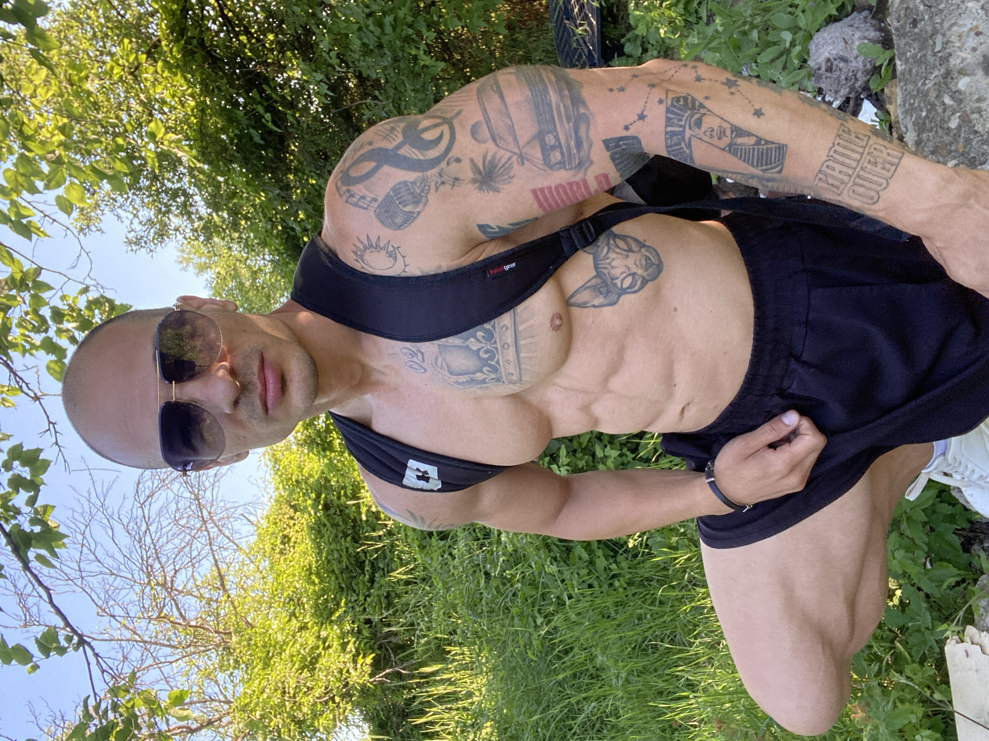 The image shows a bald, muscular man with sunglasses and a backpack. He is wearing black shorts and white sneakers. The man has numerous tattoos on his chest and arms, including a cat's face, a car, and a pharaoh. He is posing outdoors in a grassy area with trees and bushes in the background. The sky is blue and clear. There is no indication of the specific location within the city of Pleven, Bulgaria.