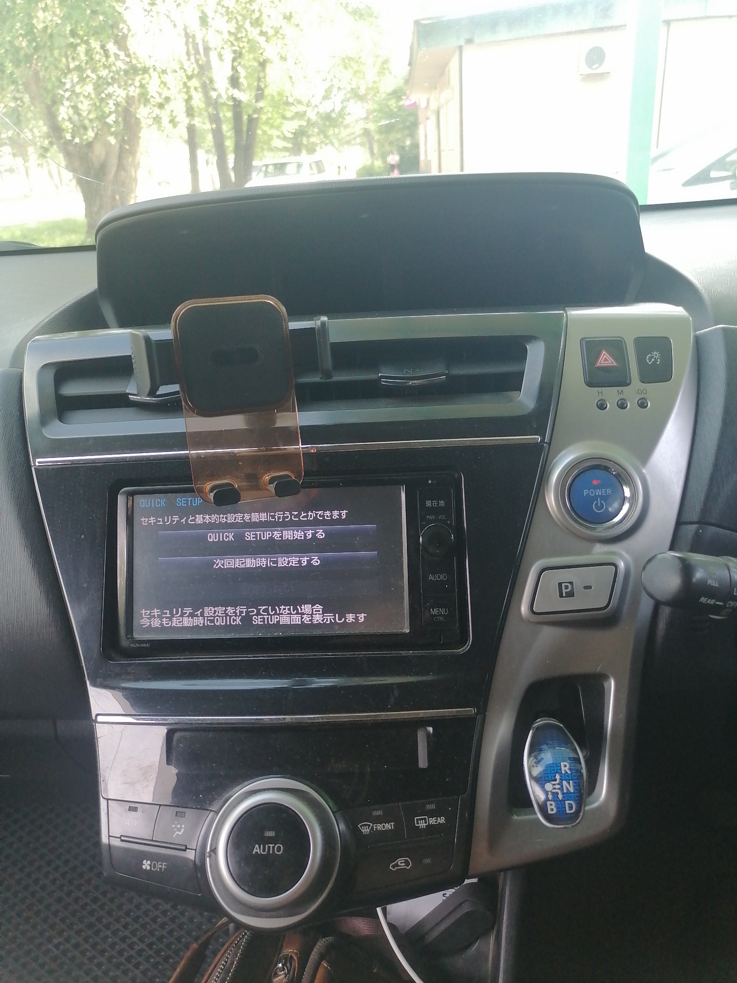 The image shows the interior of a car, likely parked outside in a sunny environment. The dashboard features a touchscreen display with Japanese text, a hazard light button, a light control dial, a blue POWER button, a parking brake button, a gear selector, and climate control buttons. A phone mount is attached to the dashboard. The exterior view through the windshield shows trees and a building. The presence of a Japanese interface on the car's electronics suggests the car may be a Japanese import or manufactured for the Japanese market. No people are visible in the image. The image itself was taken from the driver's perspective, looking at the center console and dashboard of the car.
