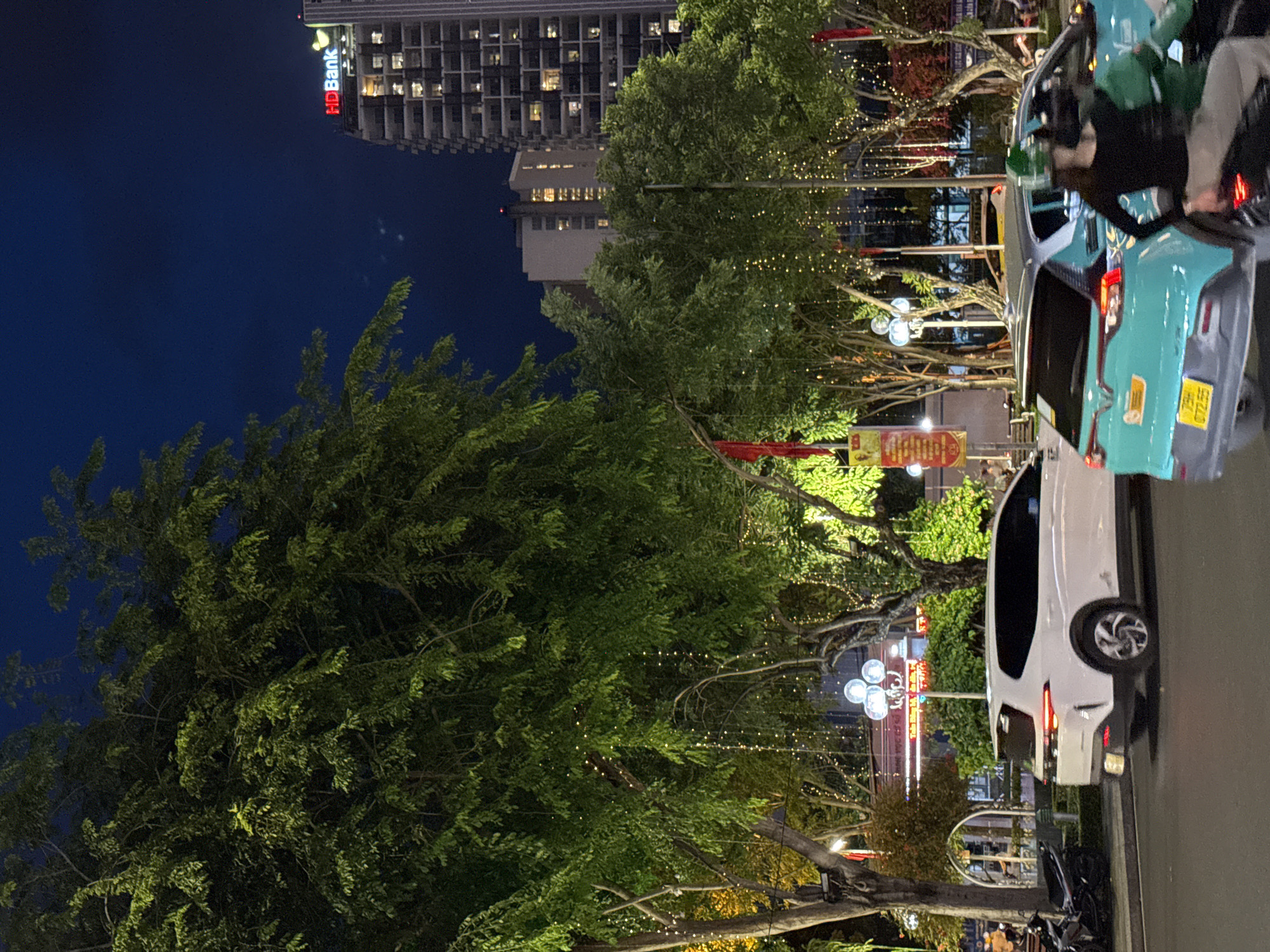 This image captures a nighttime street scene in Nha Trang, Vietnam, likely in an urban area with significant commercial activity. A large, leafy tree with decorative lights dominates the left side of the frame, its branches extending across the dark blue sky. To the right, a modern, multi-story building with illuminated windows stands tall, bearing the red "HDBank" logo and a yellow duck symbol near its peak.
The street is bustling with traffic. In the foreground, a white SUV is parked on the right side of the road, its taillights illuminated. To its right, a turquoise car, possibly a taxi, is in motion, with its rear lights visible. A person wearing a green helmet is riding a motorcycle behind the turquoise car. Further back, other vehicles and the lights of businesses are visible. The presence of decorative lights on the trees suggests a festive or commercial area, potentially a shopping district or a location for events. The overall atmosphere is that of a vibrant city at night.