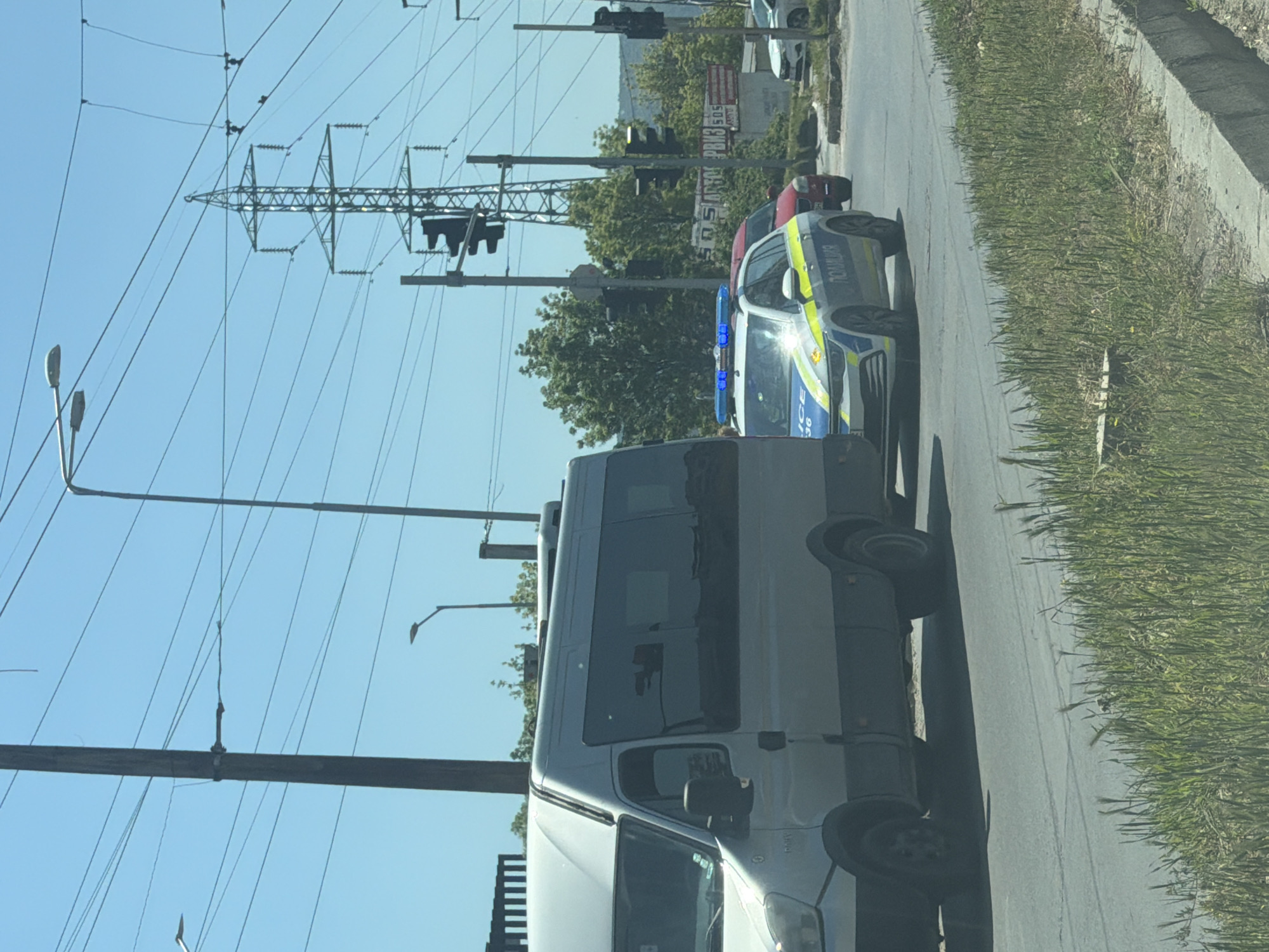 The image depicts a street scene in Varna, Bulgaria, featuring a white van and a police car parked on the roadside. Overhead, power lines run parallel to the street. Traffic lights are visible in the background, along with some buildings. A patch of green grass borders the road. The sky is clear and blue, suggesting a sunny day.
