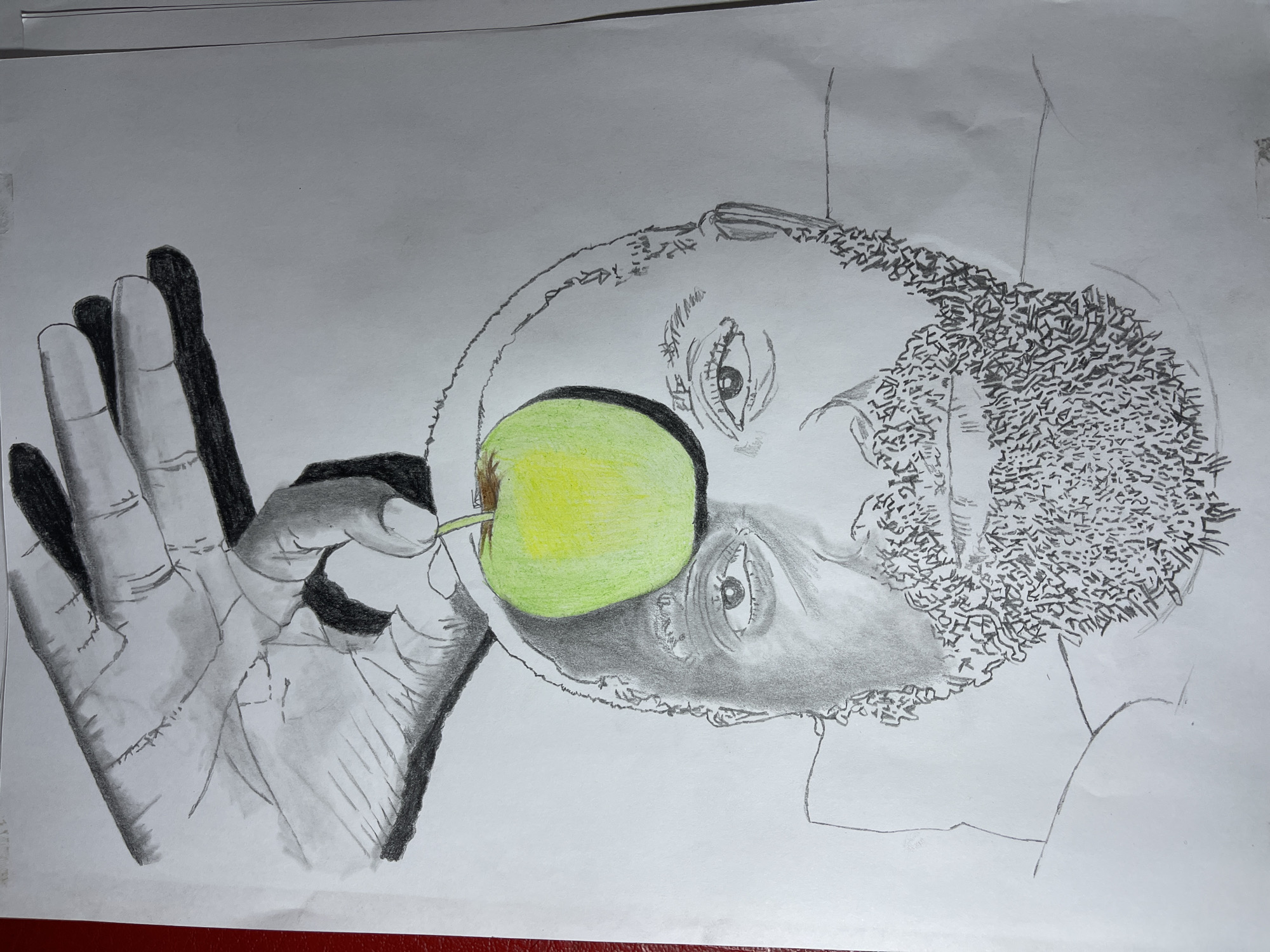 A drawing depicts a person, likely male, in profile view with an apple held in front of their face, partially obscuring it. The person's face is detailed with shading, rendering the facial features clearly, with attention paid to the eyes, nose, and mouth area. The hair is represented with a pattern of dense, short strokes, suggesting a textured or curly hairstyle. A hand, rendered in grayscale with detailed shading to show the contours and folds, is positioned holding the apple. The apple itself is colored green and yellow. There are no indications of the location being Jos, Nigeria.
