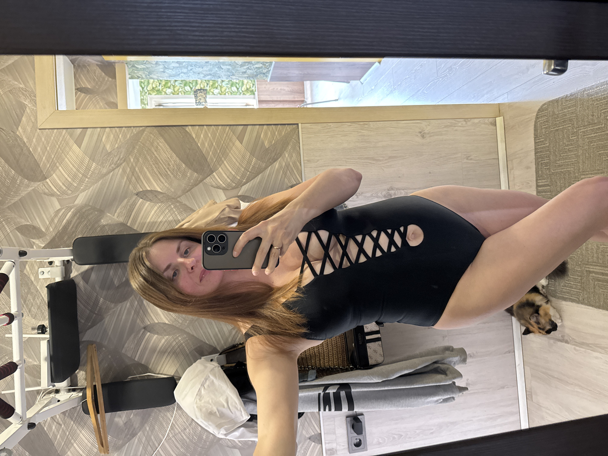 The image depicts a woman taking a selfie in a room, possibly her home, in Konakovo, Russia. She is wearing a black one-piece swimsuit with a laced cutout detail on the front. She is holding a smartphone in her right hand, facing the mirror. The room appears to be a living space or a dressing area, with a wall-mounted exercise bar visible in the background. Clothes are hanging on a rack to the left of the woman. A cat is lying on the floor near her feet. The overall lighting suggests it's daytime. The background shows a doorway leading to another room with patterned wallpaper.