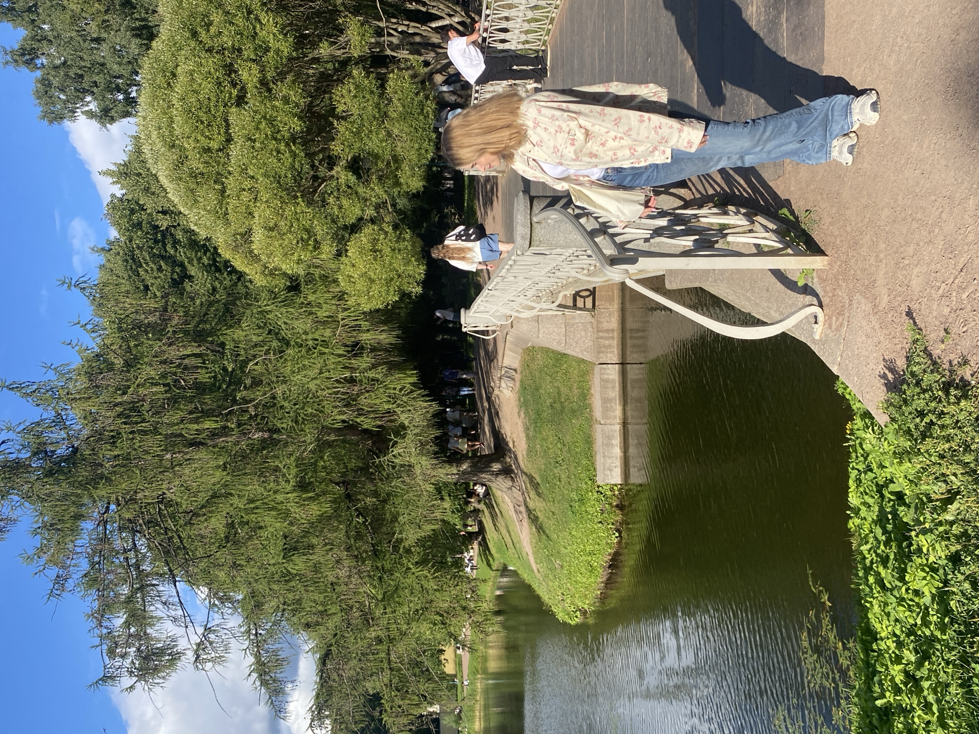 In Saint Petersburg, Russia, a young woman with blonde hair stands on a bridge overlooking a body of water. She is wearing a floral-patterned jacket, blue jeans, and white sneakers, and is carrying a light-colored tote bag. The bridge has white railings and a wooden walkway.
In the background, several other people are visible walking along a path lined with trees and grass. The scene suggests a park or garden setting, characterized by lush greenery, a serene body of water, and a bright, sunny sky with scattered clouds. The architecture of the bridge and the surrounding landscape are consistent with the style found in many of Saint Petersburg's historical parks.