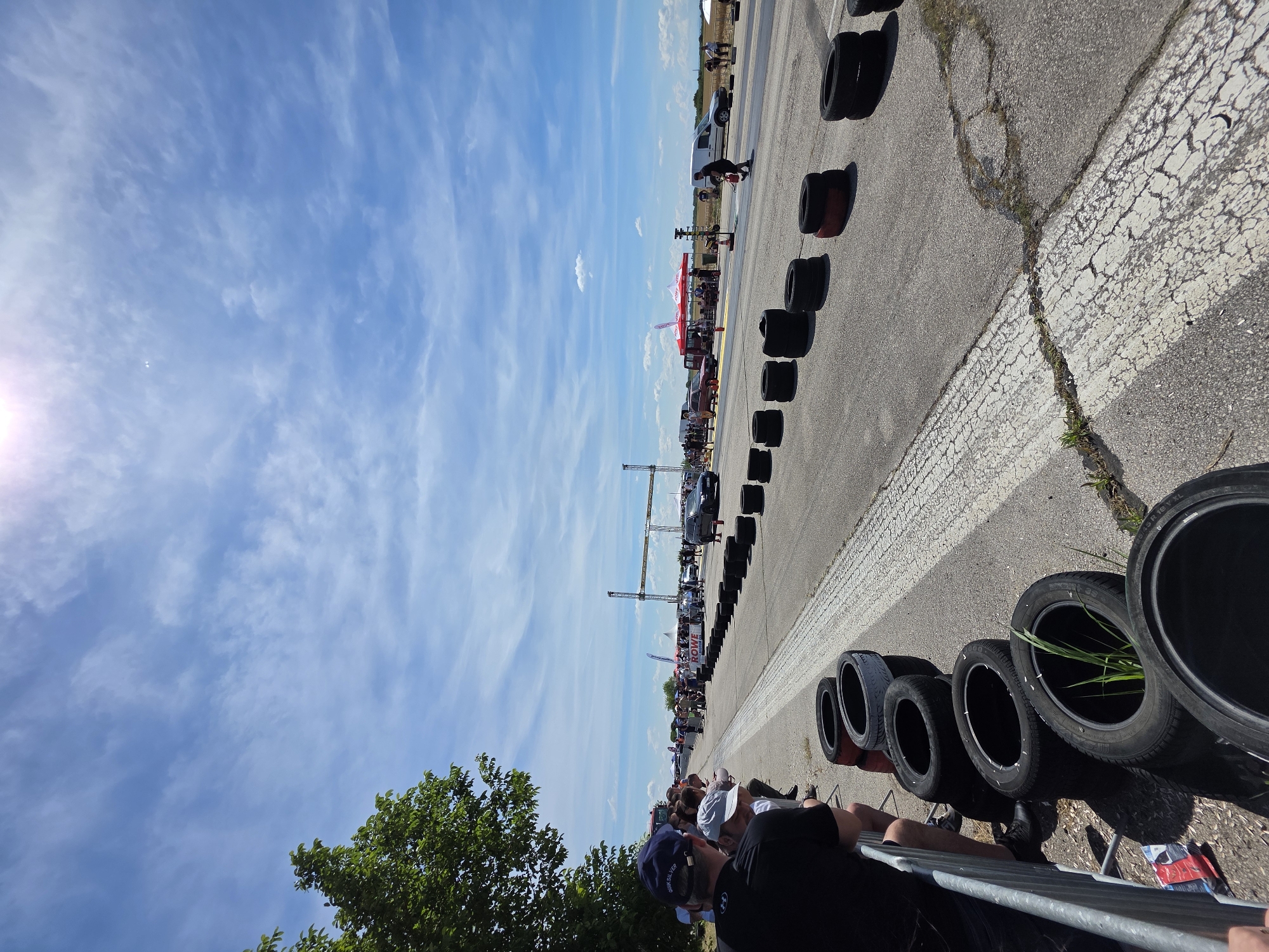 The image depicts a drag racing event taking place on a sunny day. A drag strip stretches into the distance, bordered by stacks of tires on either side. Spectators, dressed in casual attire, are visible along the left side of the track, some standing and others seated behind a metal barrier. In the background, tents and various vehicles are set up, suggesting an organized event. The sky is predominantly blue with scattered white clouds, and the sun is bright, indicating clear weather. The scene is consistent with an outdoor motorsport event.