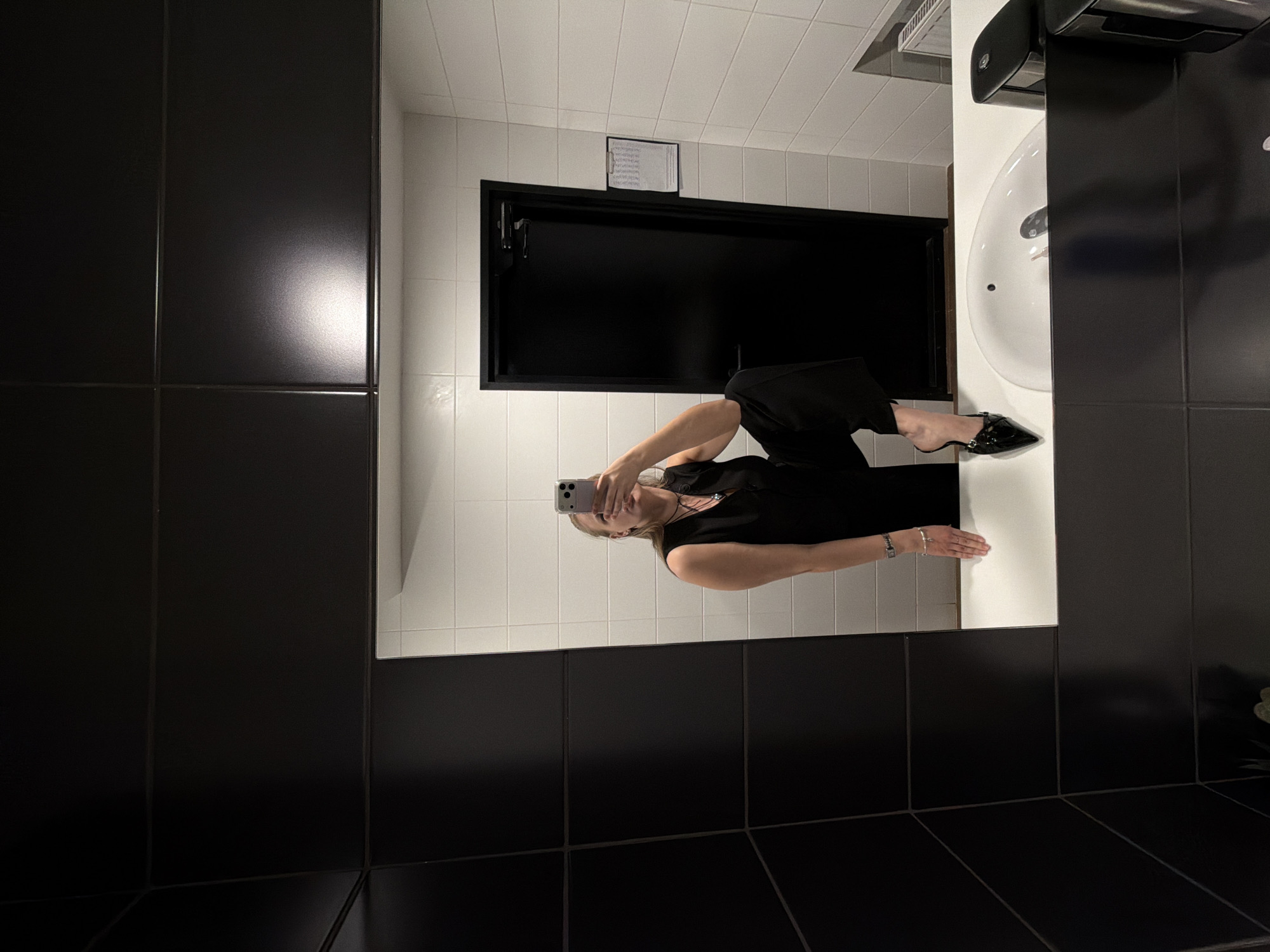 A woman is taking a selfie in a bathroom mirror. The bathroom has white tiled walls and black tiled walls. The woman is wearing a black outfit and black shoes. She is holding a phone in her right hand. The bathroom has a black door, a white sink, and a paper towel dispenser. The location and the city are not evident in the image.