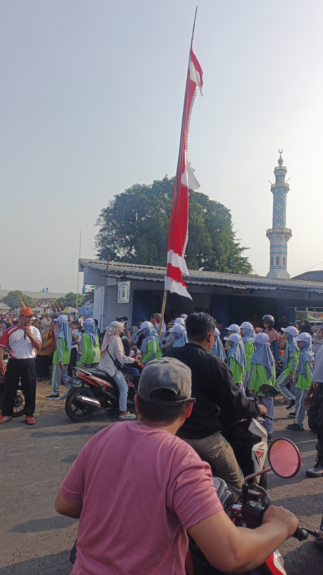 The image depicts a daytime outdoor scene, likely a public gathering or procession, in the city of Puri, Indonesia. Dominating the foreground is the back of a person wearing a pink t-shirt and a gray baseball cap, seated on a red and white motorcycle. The motorcycle's side mirror, with a pink tint, is visible. To the left and slightly behind this individual, another person in a black shirt and gray pants is also seated on a motorcycle.
In the middle ground, a crowd of people are gathered. Some individuals are wearing light blue and green uniforms with matching headscarves, suggesting they might be students or participants in an organized event. Several people are riding on motorcycles, while others are standing. A red and white flag is prominently displayed on a tall pole, extending upwards into the clear sky. Behind the crowd, a building with a simple facade and a blue awning is visible. Further in the background, a tall, slender minaret of a mosque, with white and light blue decorative bands and a crescent moon on top, is clearly discernible against the sky. A large, leafy tree stands between the building and the minaret. The ground appears to be paved. The overall atmosphere suggests a lively event under a bright, possibly hazy, sky.
