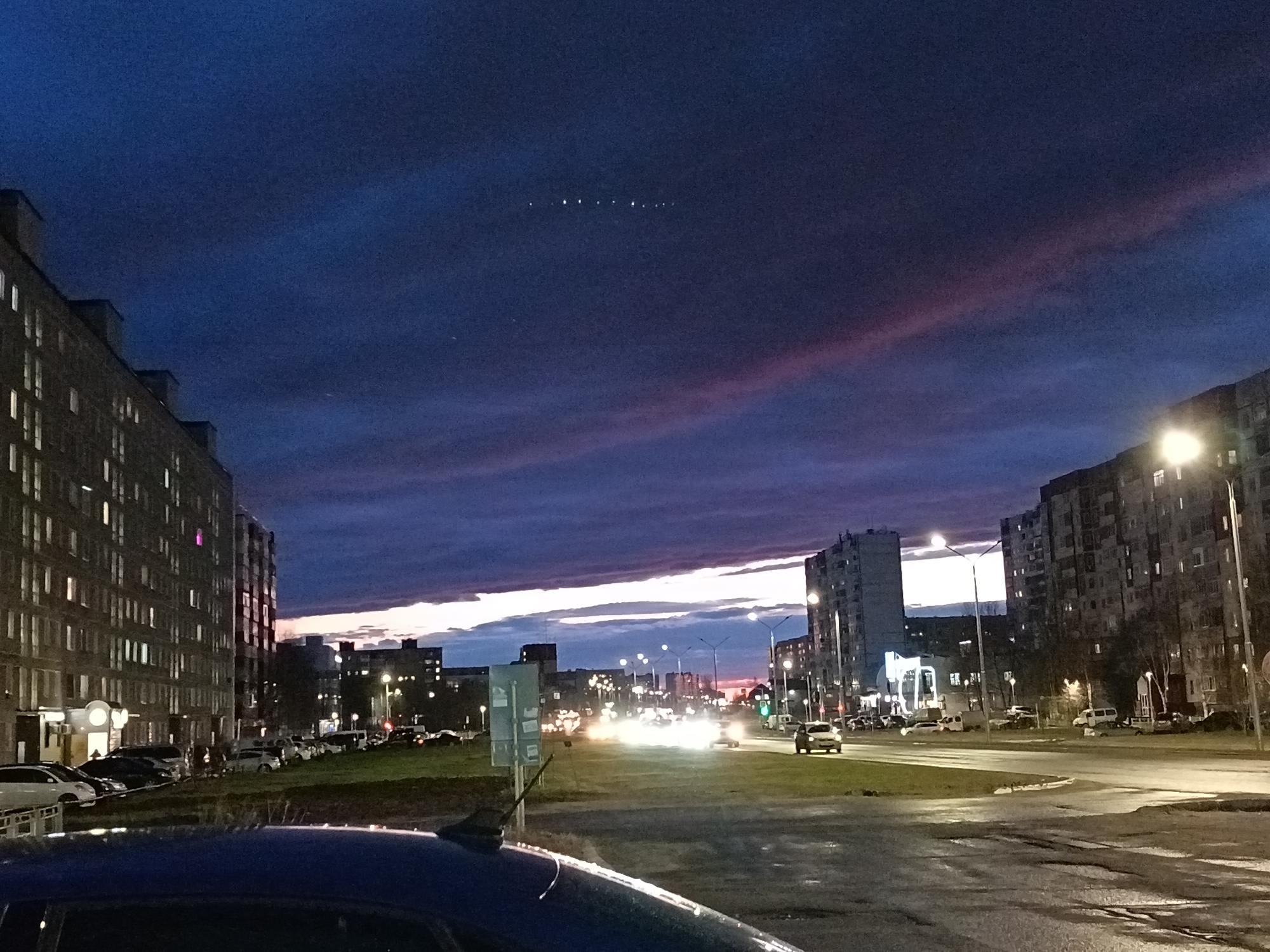 The image shows an urban scene in Nizhnevartovsk, Russia, at dusk. In the foreground is a blue car. The middle ground features a street with cars, street lights, and a traffic light. Buildings flank the street on both sides. The sky is a gradient of blue and purple, with streaks of light clouds. Above the buildings in the center of the shot are several satellites following one another in a chain across the dark blue sky.