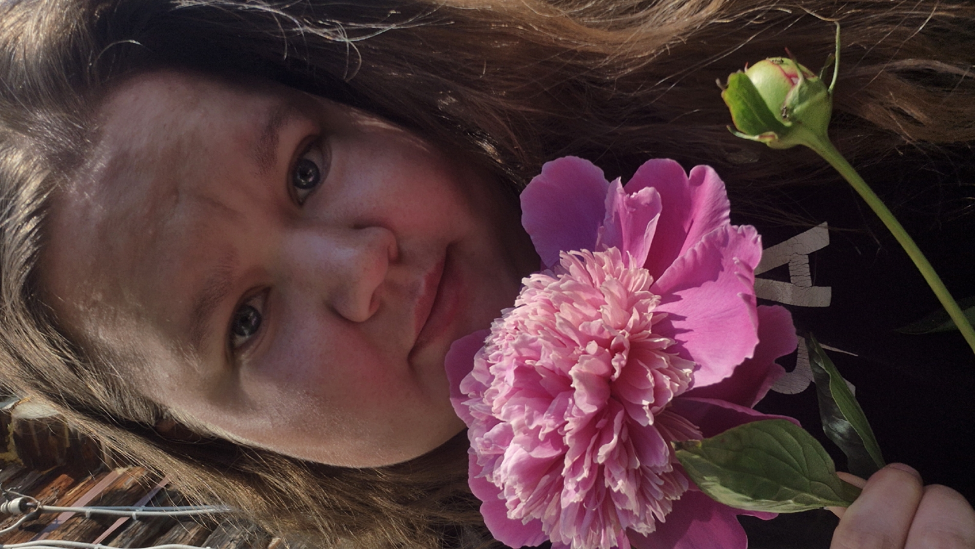 In this close-up, sunlit outdoor portrait, a person with long brown hair is holding a large, vibrant pink peony flower and its unopened bud. The person's face is partially visible, with soft features and a gentle expression. The background appears to be a rustic wooden structure, possibly a building or fence, suggesting a rural or outdoor setting. The overall mood is one of serene beauty and a connection with nature. There is no identifiable location in the city of Дойная, Russia.