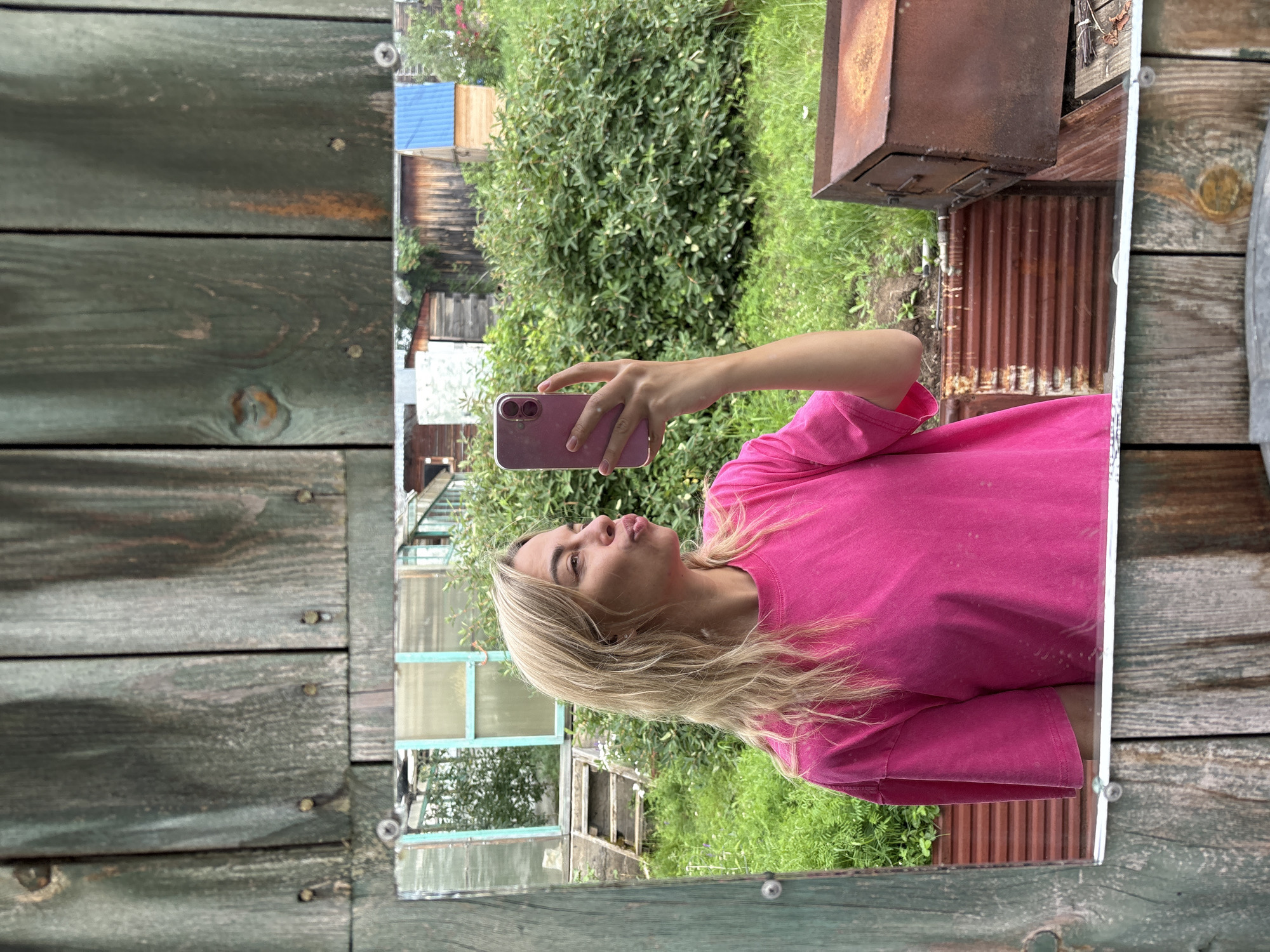 A young woman with blonde hair, wearing a pink t-shirt, takes a mirror selfie outdoors. She is holding a pink smartphone in her right hand and puckering her lips. In the reflection, she is standing in front of a wooden fence. Behind her, there is lush green vegetation, a greenhouse, and several wooden structures, including a rusted metal box that appears to be a barbecue or oven. The scene suggests a rural or backyard setting, possibly in the city of Ust-Ilimsk, Russia, given the characteristic wooden fences and structures common in such locations.