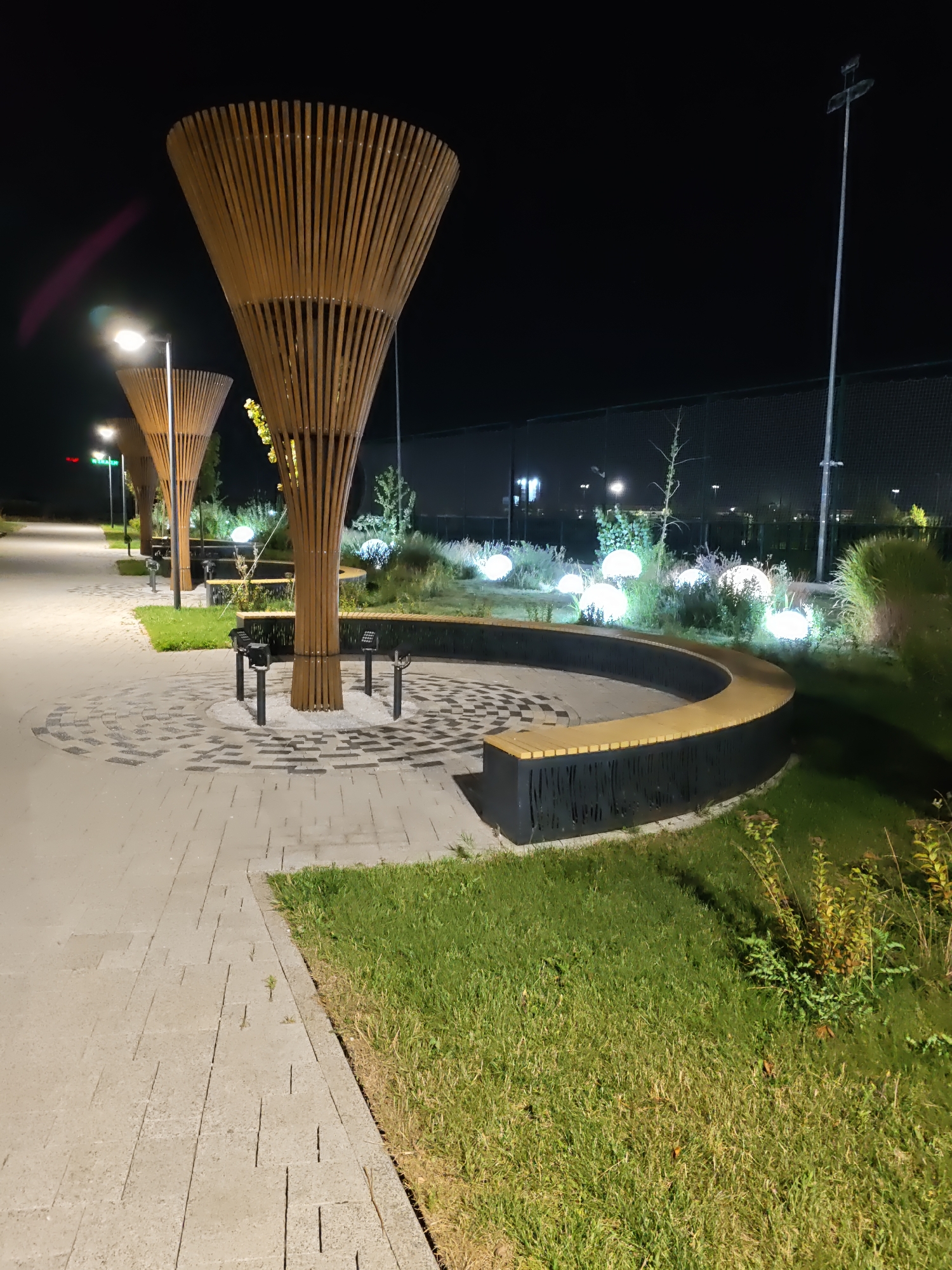 An outdoor nighttime scene captures a public park area with a paved walkway and grassy areas. The walkway is made of light-colored rectangular pavers laid in a staggered pattern, with a circular decorative paving area in the foreground featuring alternating light and dark square and rectangular tiles. Several large, cone-shaped wooden sculptures with vertical slats stand along the walkway, casting shadows. In the background, a curved bench with a dark base and light wooden seat is situated in front of a landscaped area. This landscaped area is illuminated by numerous spherical white lights scattered amongst the vegetation. Tall light poles with stadium-style lights are visible in the far background, suggesting the presence of a sports field or similar facility. The overall atmosphere is serene and modern, with the artificial lighting creating a dramatic effect against the dark night sky. There are no people or animals visible in the image.