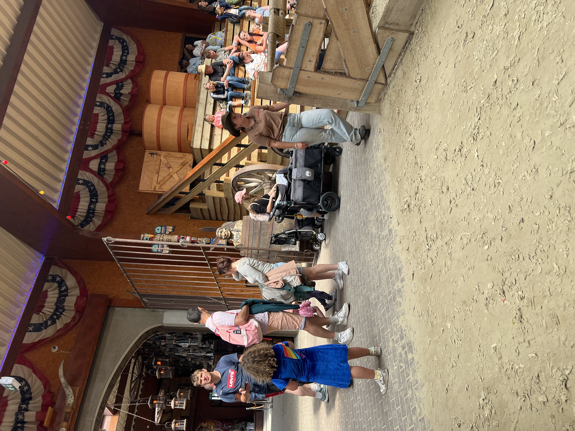 The image depicts an outdoor scene with a sandy ground and a paved area, likely in an amusement park or entertainment venue, given the rustic wooden structures, decorative banners, and presence of multiple people. In the foreground, a young girl with curly hair is wearing a blue dress with a rainbow on the back and white sneakers. To her left, a man in a blue t-shirt stands near an entrance. Behind the girl, a man in a pink shirt and shorts carries a pink backpack. A woman in a grey shirt and skirt stands to the right of the man with the backpack, holding clothing.
Further into the scene, a woman in a brown shirt, light-colored pants, and a cowboy hat walks towards the right, appearing to be an employee or performer. She is near a wooden structure and a large wagon. Several people are seated on wooden bleachers in the background, some appearing to be watching an event. Two large wooden barrels are visible behind the bleachers. The overall environment suggests a themed area, possibly with a Western or American frontier motif, indicated by the decorative elements and wooden construction. The location is not specified as Collendoorn, Netherlands in the image.