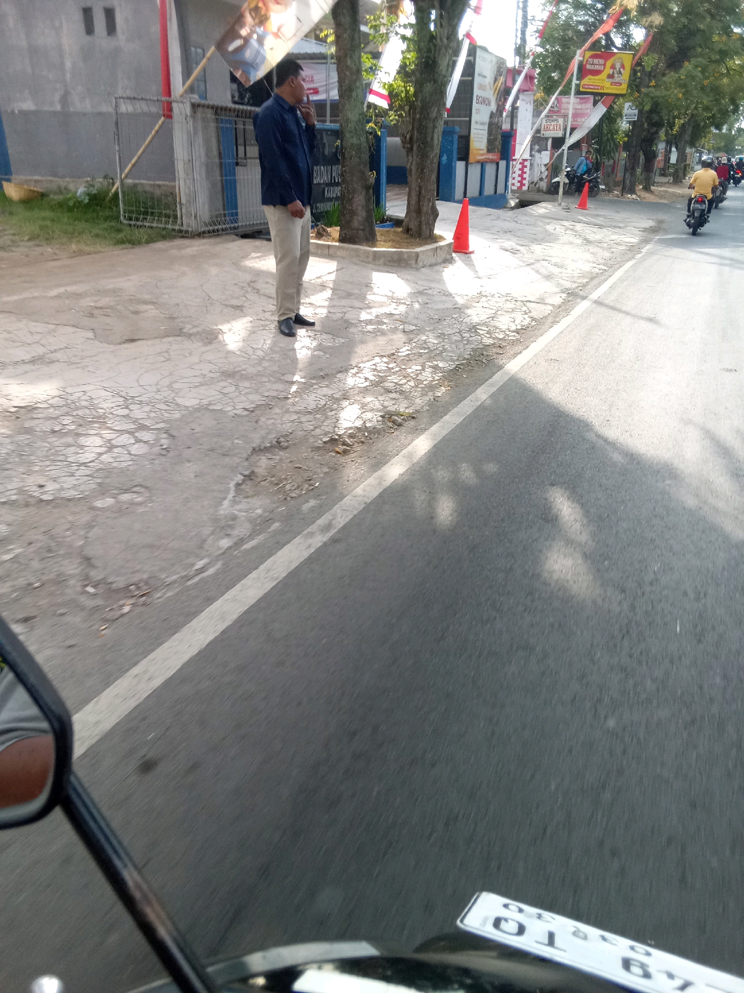 A man in a dark blue shirt and light-colored pants stands on a cracked sidewalk next to a road in Jember, Indonesia. He appears to be looking to his left, with his right hand near his face. Behind him is a building with a gray facade and barred windows, and a chain-link fence. Several trees with foliage line the sidewalk. Red and white Indonesian flags are visible, attached to poles and buildings. The road in front of him has a white line marking its edge. Several motorcycles are present on the road, with one rider in a yellow shirt visible in the distance. The scene appears to be a public street with some commercial signage visible on the buildings.