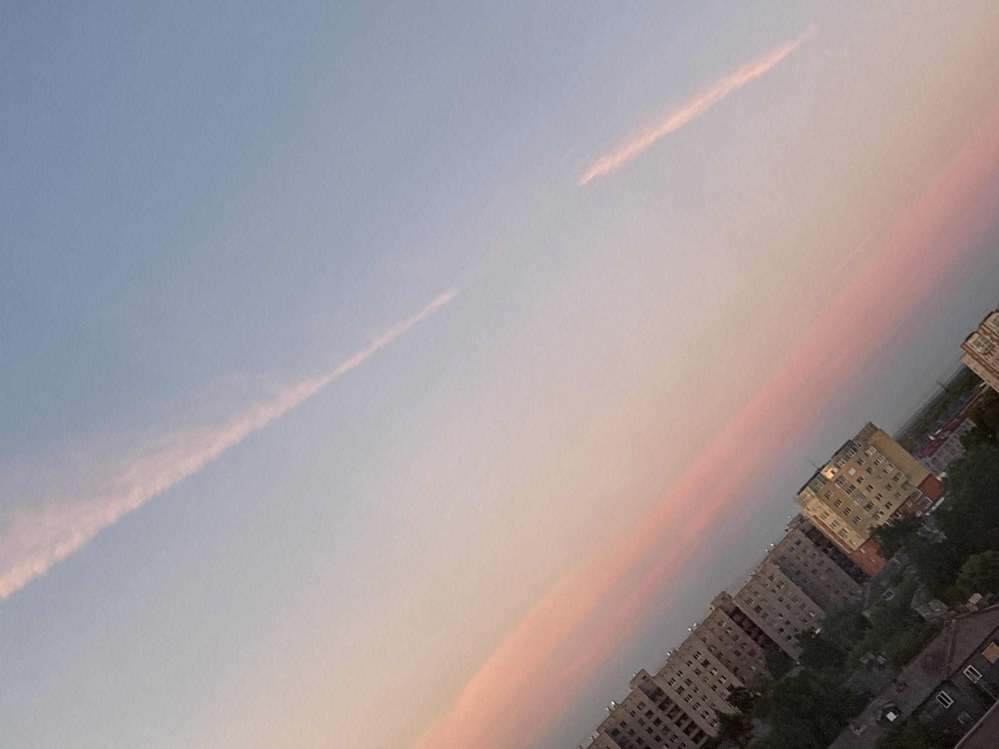 An angled shot captures apartment buildings against a pastel sky. Long, thin clouds, tinged with pink and orange, stretch across the upper portion of the frame, suggesting a sunset or sunrise. Below the sky, a cluster of apartment buildings dominates the landscape. Some are gray and appear to be multi-story structures with numerous windows. One building stands out with a yellow facade and a more distinct roofline. Green trees are visible between and around the buildings, and some smaller, darker buildings are also present in the foreground, some with visible windows. The overall scene depicts an urban environment bathed in the soft light of dawn or dusk.