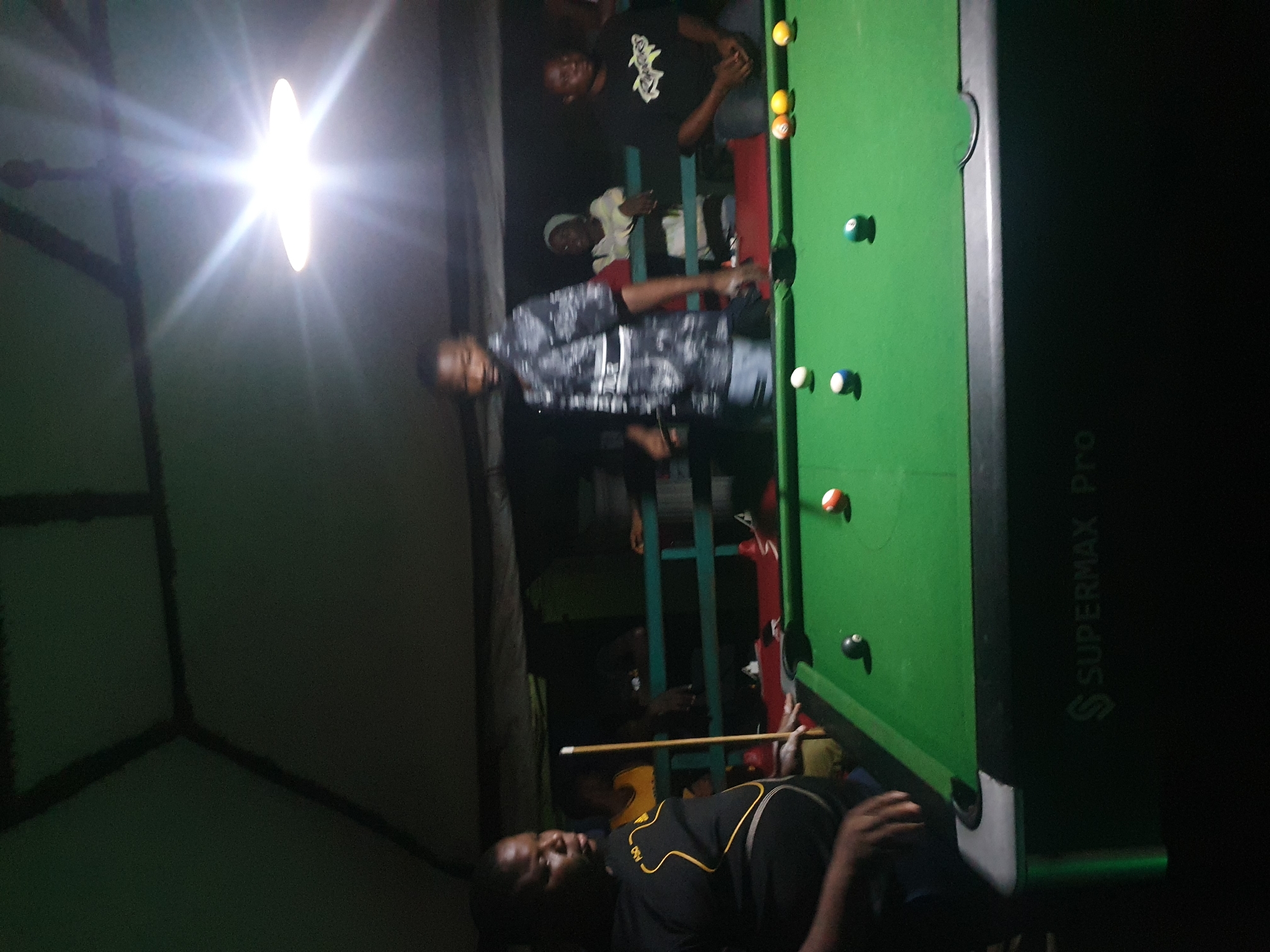This image depicts an indoor pool hall located in Lokoja, Nigeria, during the evening. A pool table with green felt is central to the scene, with several balls in play: a black 8-ball, a white cue ball, a striped blue and white ball, a solid green ball, and a solid orange ball. A pool cue rests on the table.
Three individuals are visible around the table. On the left, a person wearing a black t-shirt with a yellow design is standing and appears to be preparing to play. In the center, a person in a patterned shirt and light-colored shorts is also standing. On the right, a person in a black t-shirt is seated. In the background, other individuals are faintly visible, and a railing separates the pool area from a darker background. The environment appears to be a covered outdoor space, possibly an open-air bar or recreational area, with overhead lighting creating bright glare. The "SUPERMAX Pro" logo is visible on the side of the pool table.