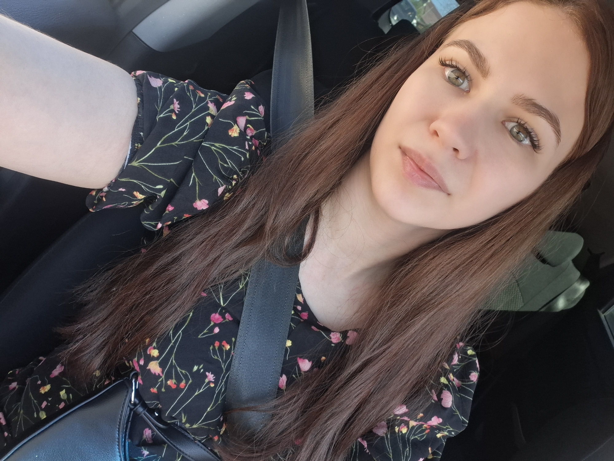 A young woman is taking a selfie in the driver's seat of a vehicle. She has long, brown hair, fair skin, and green eyes. She is wearing a black floral dress and a seatbelt. A black purse is partially visible on her lap. Based on the location information provided, it is inferred that the photo was taken somewhere in the city of Chaikovsky, Russia. However, no specific landmark or street can be identified from the image.