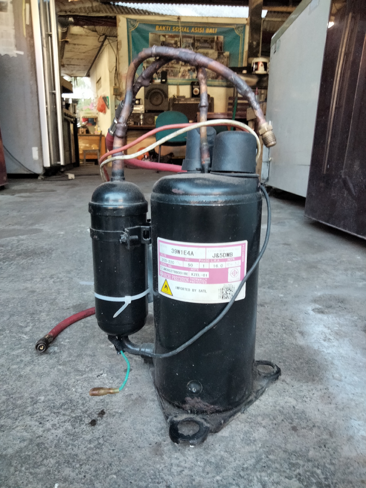 In Denpasar, Indonesia, a black compressor unit for a refrigerator or air conditioning system rests on a gray, weathered concrete floor. The compressor has a label that reads "39W1E4A" and "J&5DMB". It is connected to a smaller cylindrical component by copper tubing and wiring. A red hose with a brass fitting is attached to the side of the unit. The background shows an open-air workshop or storage area with various items including refrigerators, shelves, and a teal banner with Indonesian text. The lighting suggests an overcast day or an indoor setting with artificial light.