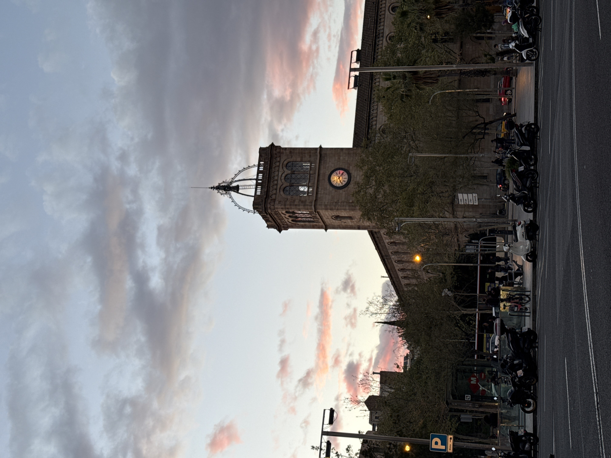 The image depicts the main building of the University of Barcelona, viewed from street level. The building's clock tower is visible against a backdrop of a cloudy sky, with some hints of pink from the setting sun. Along the street, several motorcycles are parked. There are street lights and other infrastructure present. The image seems to be taken at dusk or dawn.