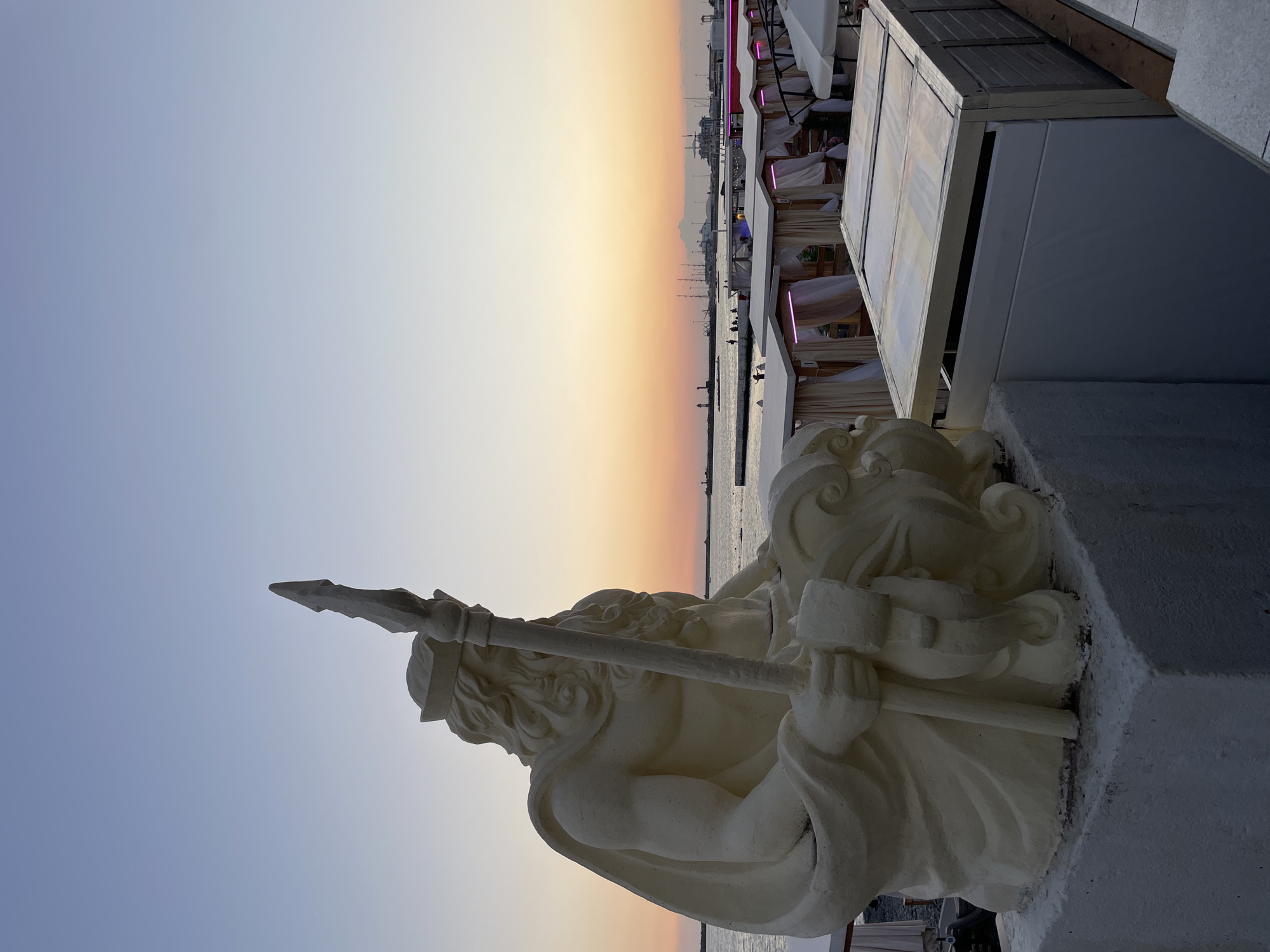 A large, cream-colored statue of a seated male figure, possibly Neptune or a similar mythological character, holds a trident and is adorned with a cape and a crown. The statue is positioned on a white pedestal overlooking the sea, with a beach lined with white cabanas with pink lighting in the background. The sky displays a gradient of sunset colors, transitioning from pale blue to orange. Several boats and a lighthouse are visible on the horizon. The overall scene suggests a resort or recreational area at dusk. This statue is located on the embankment in the city of Sochi, Russia, at the beach near the Marine Station.