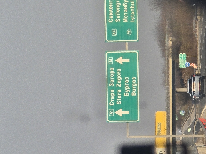 The scene depicts a multi-lane highway interchange near Cherna Gora, Bulgaria, specifically the junction of the A1 (Trakia) and A4 (Maritsa) motorways, under an overcast, light grey sky.
Two prominent green overhead gantry signs provide directional information. The left sign displays "A1" with text "Стара Загора / Stara Zagora" and "Бургас / Burgas," each with an upward arrow, indicating continuation on the A1 motorway. The right sign shows "A4" with text "Свиленград / Svilengrad" and "Истанбул / Istanbul," with a "TR" country code symbol for Turkey, indicating a diverging route.
The highway consists of dark asphalt lanes. A light-colored SUV is visible in the foreground, traveling away from the viewer on the main highway. Overhead, an elevated concrete bridge or overpass crosses perpendicular to the highway, supported by concrete pillars. A yellow truck trailer with "DÜZTAS" printed on its side is seen moving on this overpass. Sparse, bare trees and bushes line the highway shoulders and areas beneath the overpass, consistent with late autumn or winter vegetation. Smaller green and blue directional signs, along with red and white traffic barriers, are visible at ground level near the road. No individuals are explicitly visible.