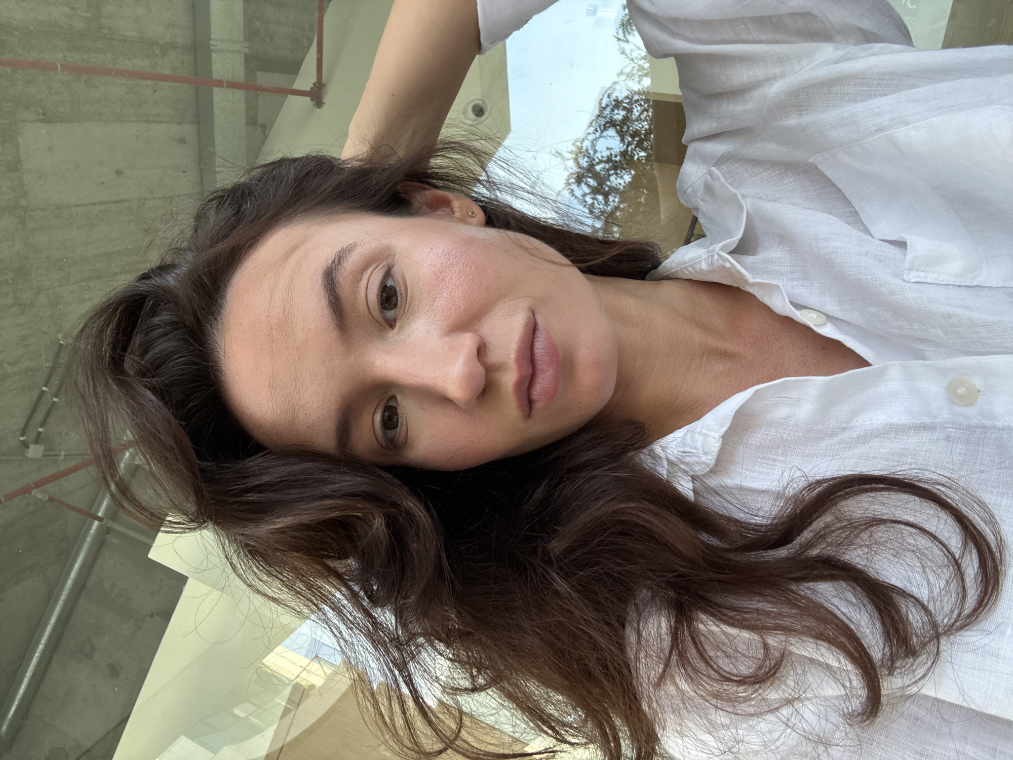 A portrait of a woman with long, dark, wavy hair and light skin. She is wearing a white linen shirt. Her right hand is resting on her head, and her left arm is down. The background appears to be indoors, possibly a cafe or office space, with exposed concrete ceilings and pipes, suggesting an industrial or modern architectural style. Through a large window, greenery and trees are visible, hinting at an outdoor environment. The overall lighting suggests daylight. The image composition is a close-up selfie.
