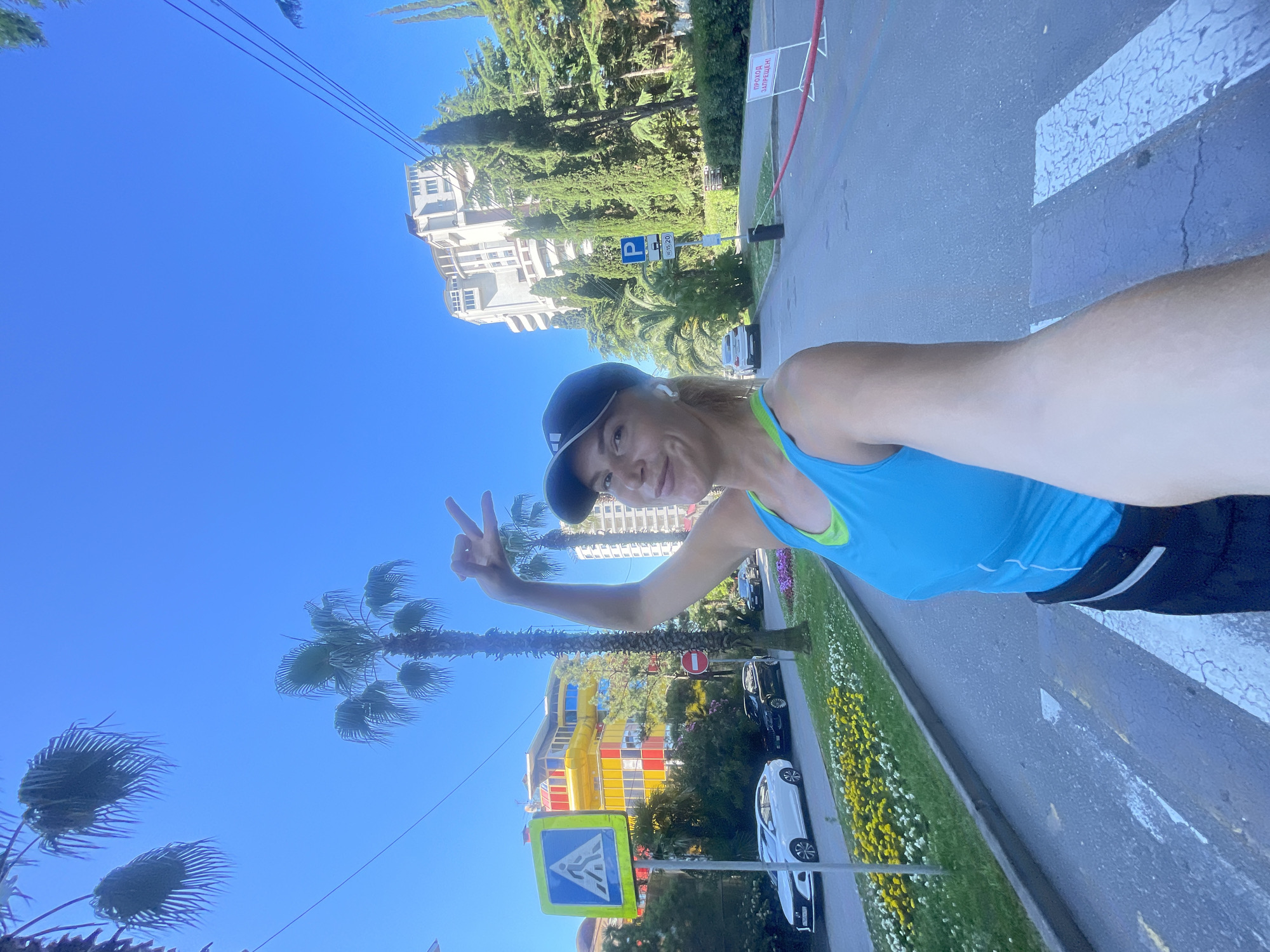In Sochi, Russia, a woman in athletic wear takes a selfie while standing at a pedestrian crossing. She wears a blue tank top, a black baseball cap, and white earbuds. Her right hand is raised, forming a peace sign. Behind her, palm trees and cypress trees line a street with a sidewalk adorned with flowers. Buildings with colorful facades and a multi-story hotel are visible in the background. Several cars are parked along the street, and traffic signs, including a pedestrian crossing sign and a parking sign, are present. A red barrier tape with Russian text that translates to "ENTRY PROHIBITED" is also visible. The sky is clear and blue.
