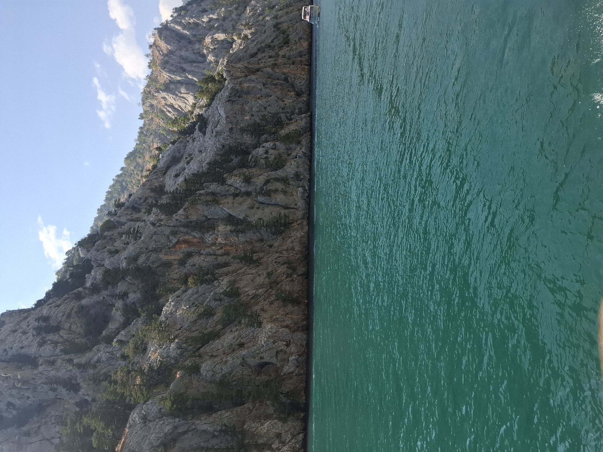 A boat is traveling through a turquoise body of water, with steep, rocky cliffs rising on the left side of the frame. The cliffs are covered in sparse vegetation, primarily small trees and bushes. The sky is blue with a few scattered white clouds. The boat is white with a red stripe and is moving away from the viewer towards the right side of the image. The scene is likely a canyon or a gorge, possibly the Oymapinar Dam or a similar natural formation near Manavgat, Turkey, known for its scenic beauty and water activities.