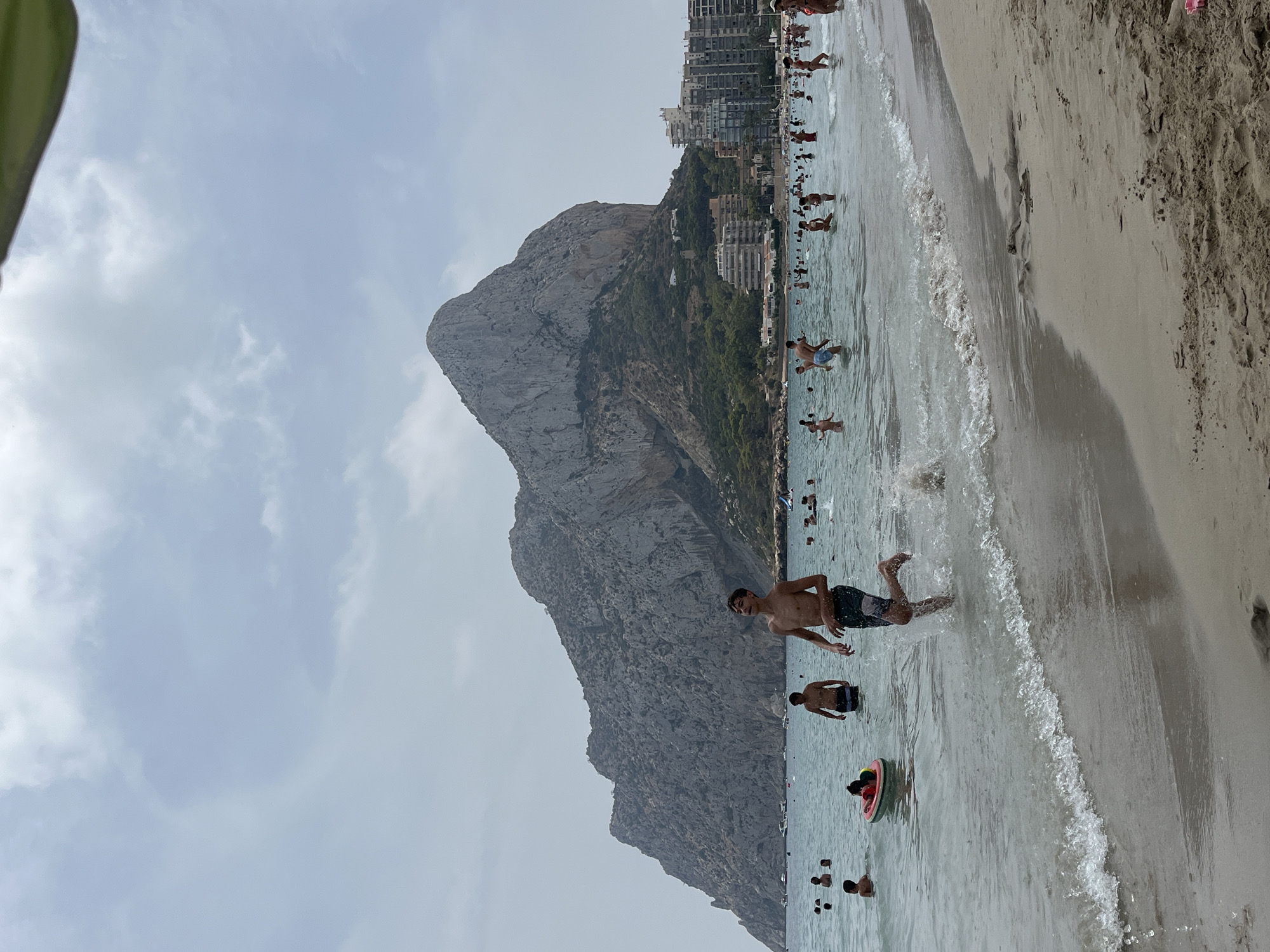 This is a photograph of a beach scene in the city of Calpe, Spain. In the foreground, a young man is running through the shallow water towards the camera, creating a splash. He is wearing dark swim trunks. The water is clear and calm, with small waves breaking on the sandy shore.
In the middle ground, numerous people are enjoying the water, swimming, standing, and playing. Some are visible in the distance, further out in the sea. One person is on a small, inflatable ring. The water extends to a shoreline with buildings lining the coast.
Dominating the background is a large, rocky mountain that rises dramatically from the sea. The mountain is grey and textured, with some vegetation visible on its lower slopes. Behind the mountain, a cluster of tall, modern buildings can be seen, characteristic of a coastal city. The sky is overcast with a hazy, light grey cloud cover. The overall atmosphere is that of a busy beach on a warm, cloudy day.