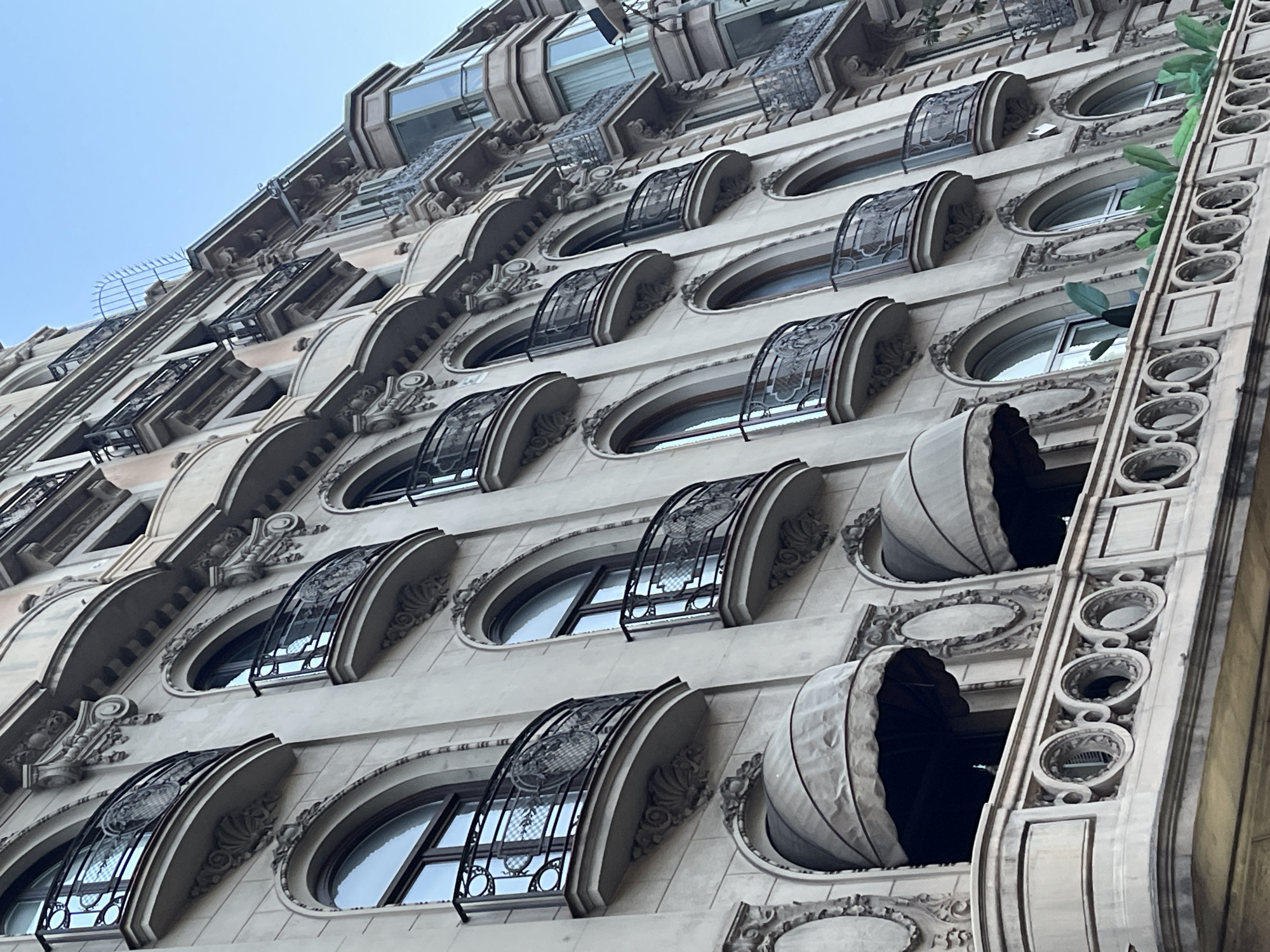 This is a close-up, low-angle shot of the exterior of a historical building in Barcelona, Spain. The building features a pale, textured facade adorned with ornate architectural details, including intricate moldings, carved embellishments around arched windows, and decorative balconies. Several arched windows with dark frames are visible, some with curved black metal railings, creating Juliet balconies. Above some of these windows are beige fabric awnings. A prominent decorative balustrade runs along a lower level, featuring a repeating pattern of circular openings. Lush green plants are seen peeking out from behind this balustrade. The sky is a clear, pale blue. The architecture suggests a period style, possibly Art Nouveau or a similar European style popular in the late 19th or early 20th century.