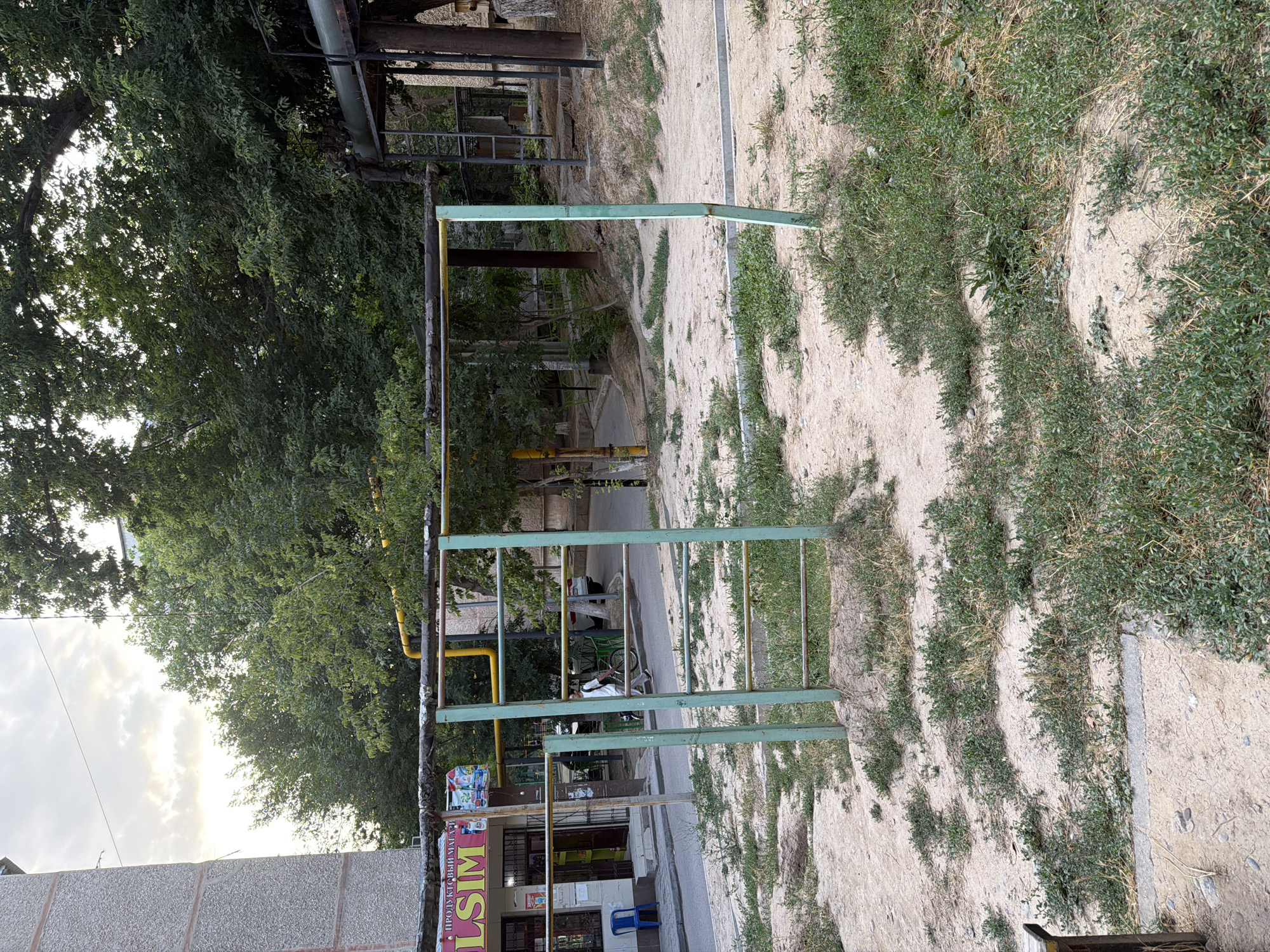 The image captures an outdoor scene on a cloudy day, likely in the city of Shymkent, Kazakhstan, judging by the architecture and vegetation. In the foreground, a weathered, light green metal playground apparatus stands on a sandy, uneven ground covered with sparse, dry grass. This structure features horizontal bars and vertical supports, with some of the bars appearing rusted.
Behind the playground, a street runs horizontally. On the left side of the street, a grey, multi-story apartment building with textured concrete walls is visible. Adjacent to the building is a small shop with a brightly colored sign, part of which reads "LSIM" in yellow lettering, suggesting it might be a grocery store or convenience shop. A blue plastic chair sits outside the shop entrance.
Further down the street, a person wearing a white shirt and dark pants is walking while pushing a bicycle. A white car is parked further down the road. The background is dominated by lush green trees, their branches extending into the frame, some obscuring parts of buildings. Industrial-looking pipes, one with a yellow coating, are also visible among the trees and structures, hinting at the surrounding urban infrastructure. The overall impression is that of a residential area with aging public amenities.