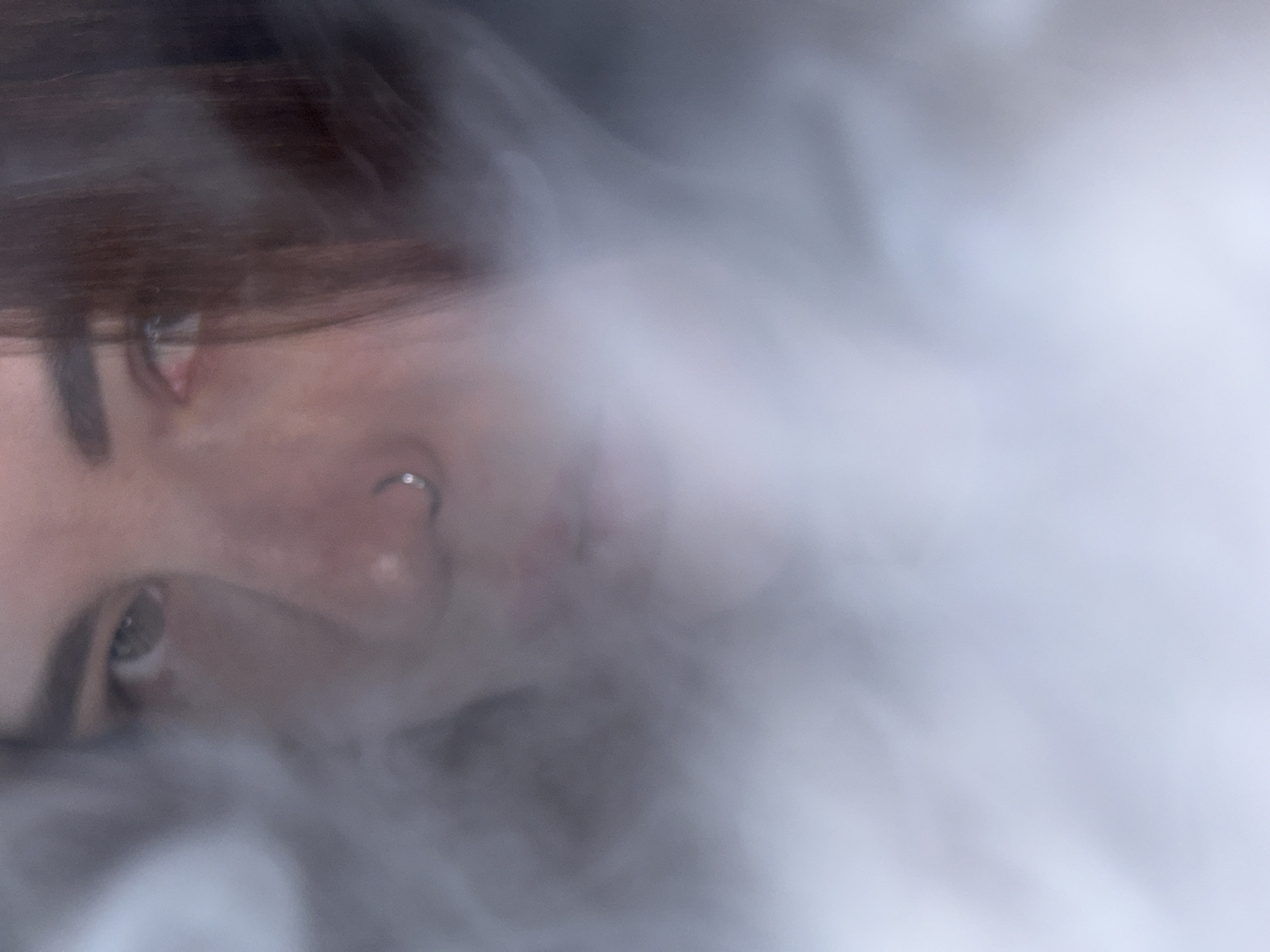 A close-up shot of a person's face obscured by smoke or fog. Visible features include the left eye, eyebrow, nose with a piercing, and dark hair. The background is out of focus, consisting mostly of the indistinct smoke. The location is not discernible from the image.