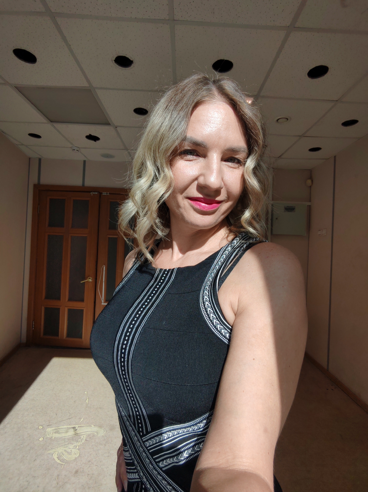A woman with blonde, wavy hair and fair skin is centered in the frame, facing the camera with a slight smile. She is wearing a black, sleeveless dress with white patterned accents along the straps and torso. The photo appears to be taken indoors in a hallway. Behind the woman are double wooden doors with glass panels, and a beige wall. The ceiling is a standard grid pattern with recessed lighting fixtures. There's sunlight casting shadows on the walls and floor, giving the scene a warm tone. There is no information on the city and location.