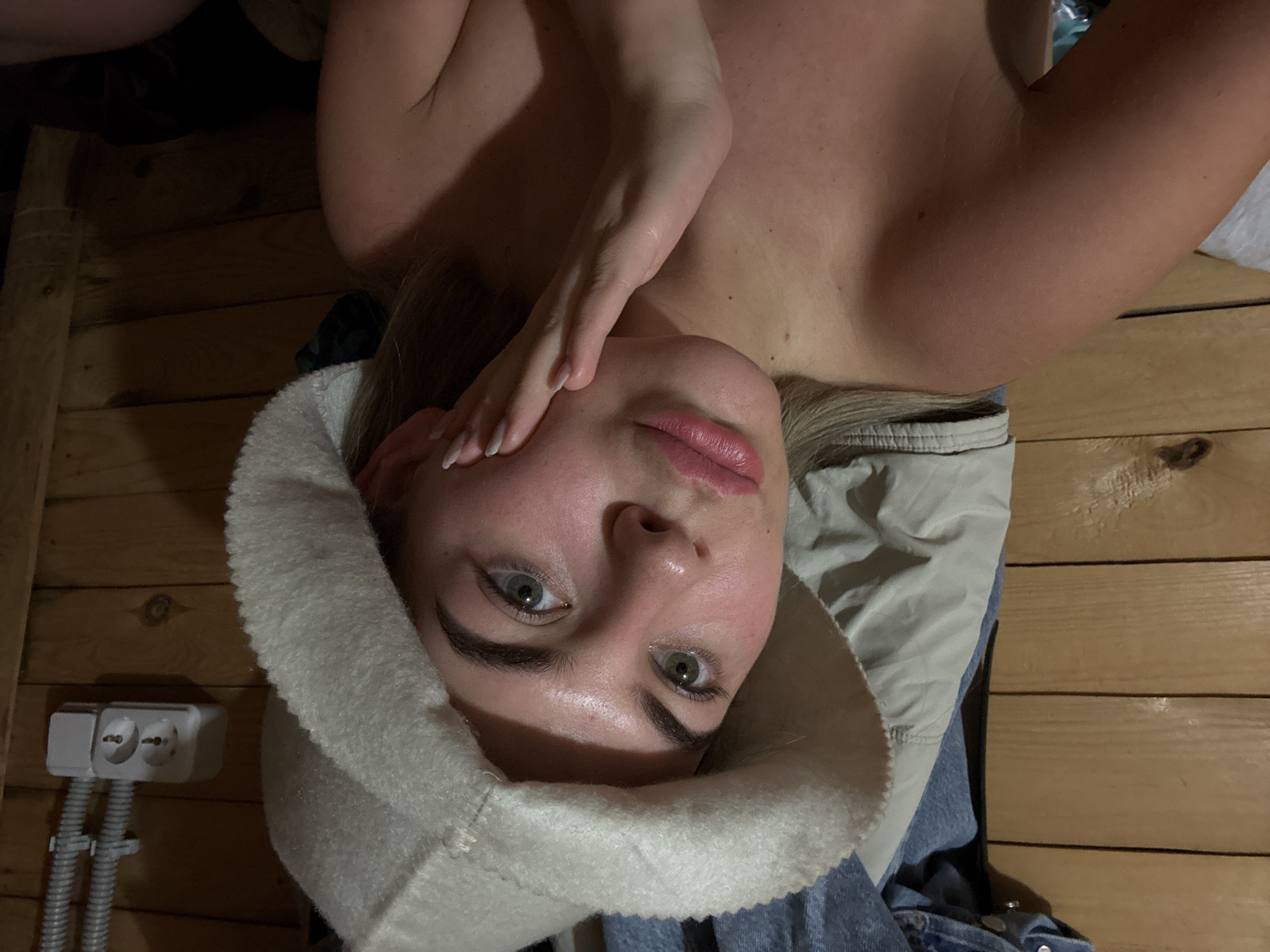 The image captures an overhead view of a person, likely female, lying down and looking directly at the camera. The subject is oriented with her head towards the bottom of the frame and her body extending upwards.
She is wearing a fluffy, light-colored, possibly white or cream-colored, hat that covers her head and ears, resembling a traditional sauna hat. Her face is clearly visible, featuring green or hazel eyes, dark eyebrows, and full lips with a hint of pink lipstick. Her skin appears fair with some natural flush. One of her hands is gently placed on the side of her face, near her temple/cheek, with manicured nails that have white tips.
Her upper body appears bare or topless, revealing her shoulders and chest. Around her, various fabrics are visible: a light gray or beige material, possibly a blanket or jacket, and blue denim, suggesting jeans or another piece of clothing.
The setting appears to be an indoor space with wooden plank walls, arranged horizontally. To the left side of the frame, mounted on the wooden wall, is a white electrical outlet featuring two European-style sockets. Two grey corrugated conduits run vertically downwards from the outlet. The overall lighting is somewhat dim, suggesting an intimate or relaxed atmosphere, possibly indoors at any time of day, with artificial light sources.
The presence of the sauna hat and the wooden interior strongly suggest the person is in a sauna, steam room, or a similar relaxation area, likely in a moment of rest or contemplation. The direct gaze into the camera implies a personal photograph, possibly a selfie. There is no visible text in the image.