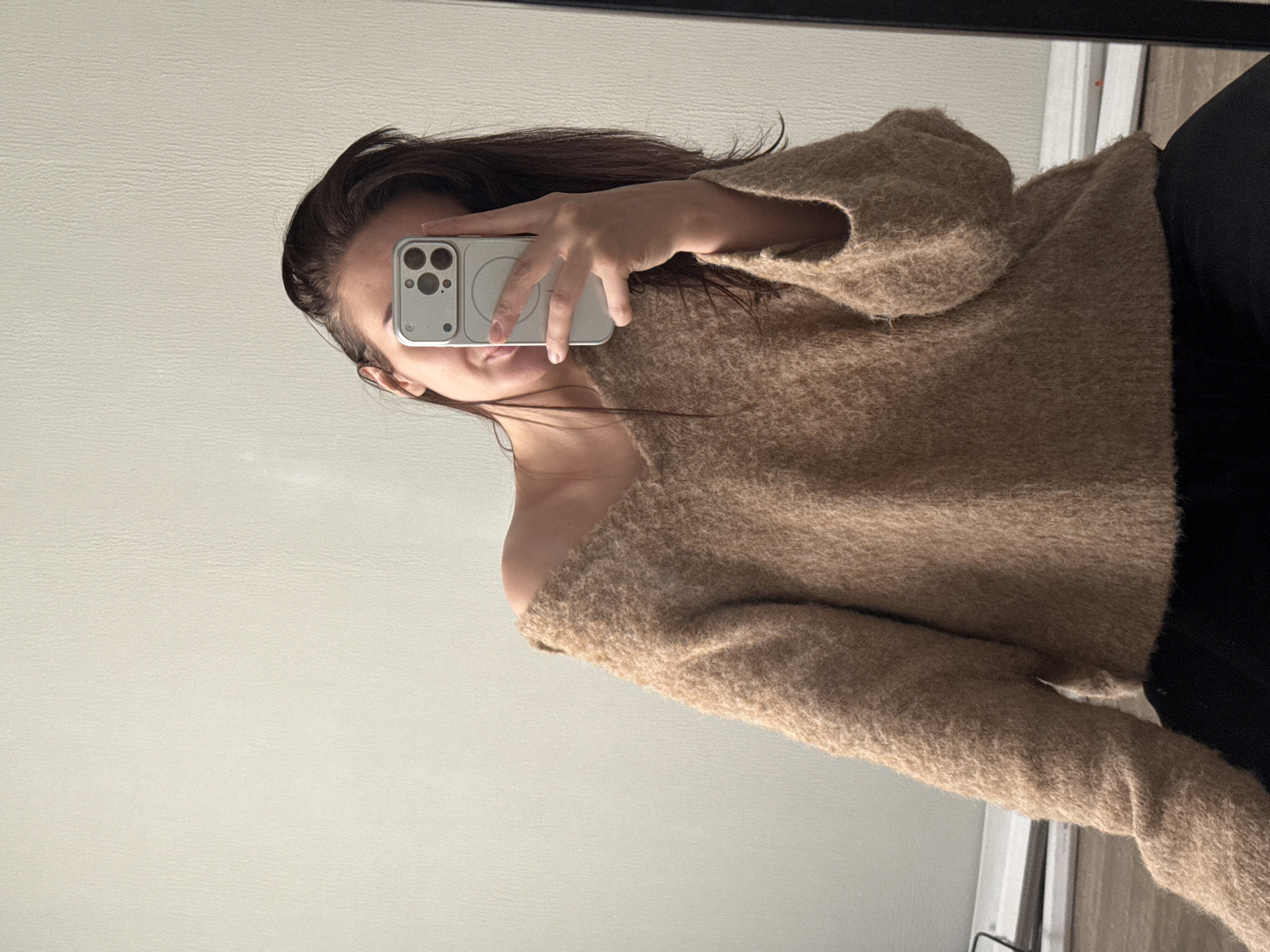 A woman is taking a selfie in a mirror. She is wearing a brown sweater that is off-the-shoulder on one side and black pants. The background shows a plain wall. There is no indication of a specific location such as Chaikovsky, Russia.