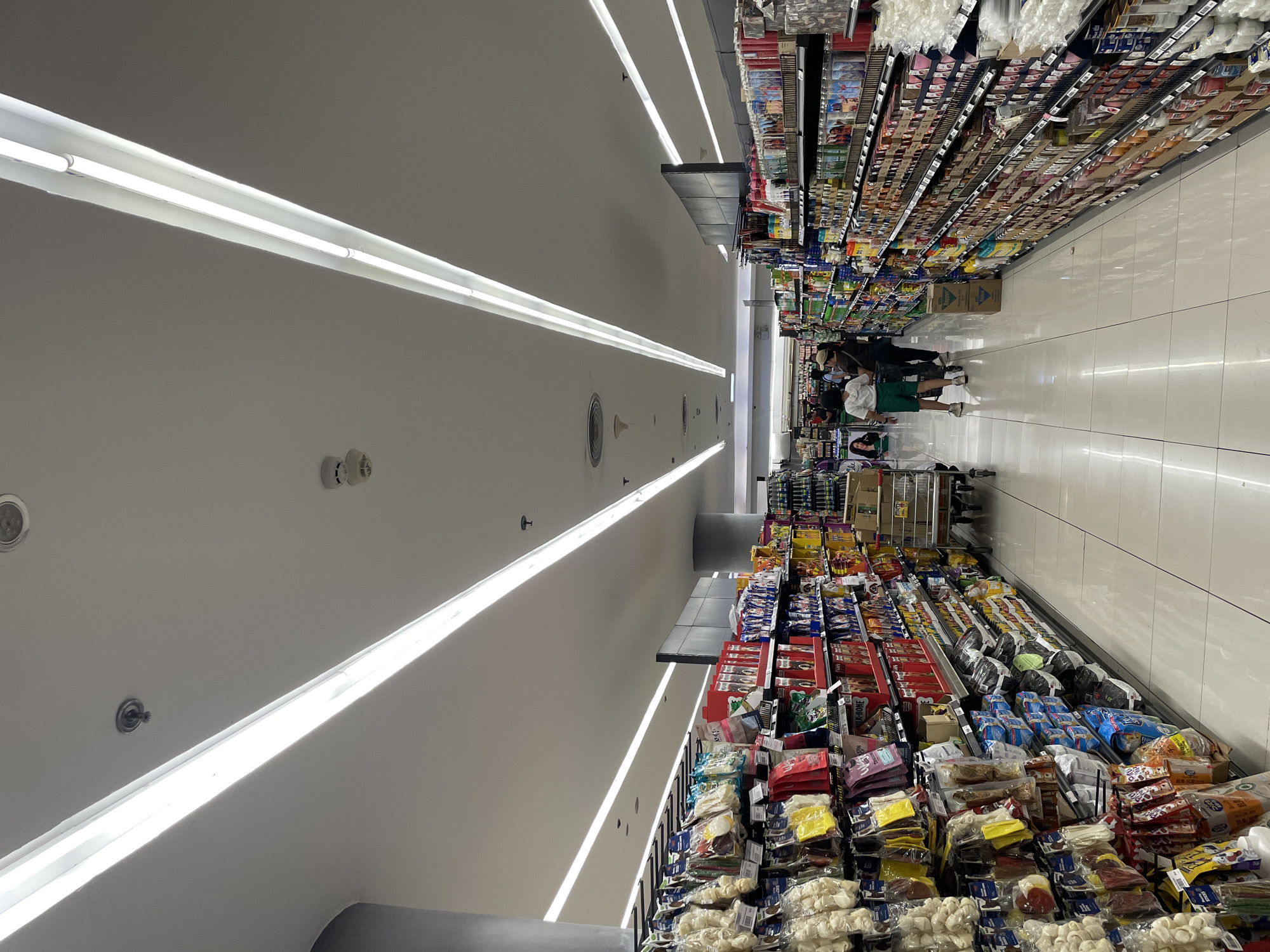 A brightly lit, long aisle in a supermarket stretches into the distance, lined with shelves stocked with various products, predominantly pet supplies on the left and household goods or toiletries on the right. The floor is a clean, white tiled surface. In the middle distance, three individuals are visible walking away from the camera. The aisle is illuminated by long, rectangular fluorescent light fixtures running along the ceiling. The scene appears to be a typical retail environment within a commercial establishment. This image was captured in Davao City, Philippines.