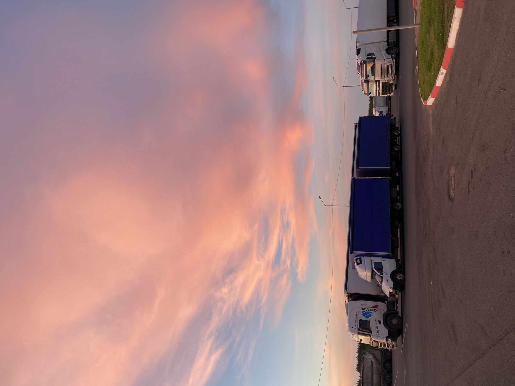 The image depicts a parking lot scene at dusk, characterized by a vibrant pink and blue sky filled with wispy clouds. Several semi-trailer trucks are parked in the lot. In the foreground, a white semi-truck with a blue trailer is parked on the right, and to its left is another white semi-truck with a blue trailer, and a smaller white delivery van in front of it. Further to the left, another white semi-truck is visible. The ground is asphalt, with a red and white curb bordering a patch of grass on the right. Streetlights are visible in the background. The location is identified as Sazonovo, Russia. There are no people visible in the image.