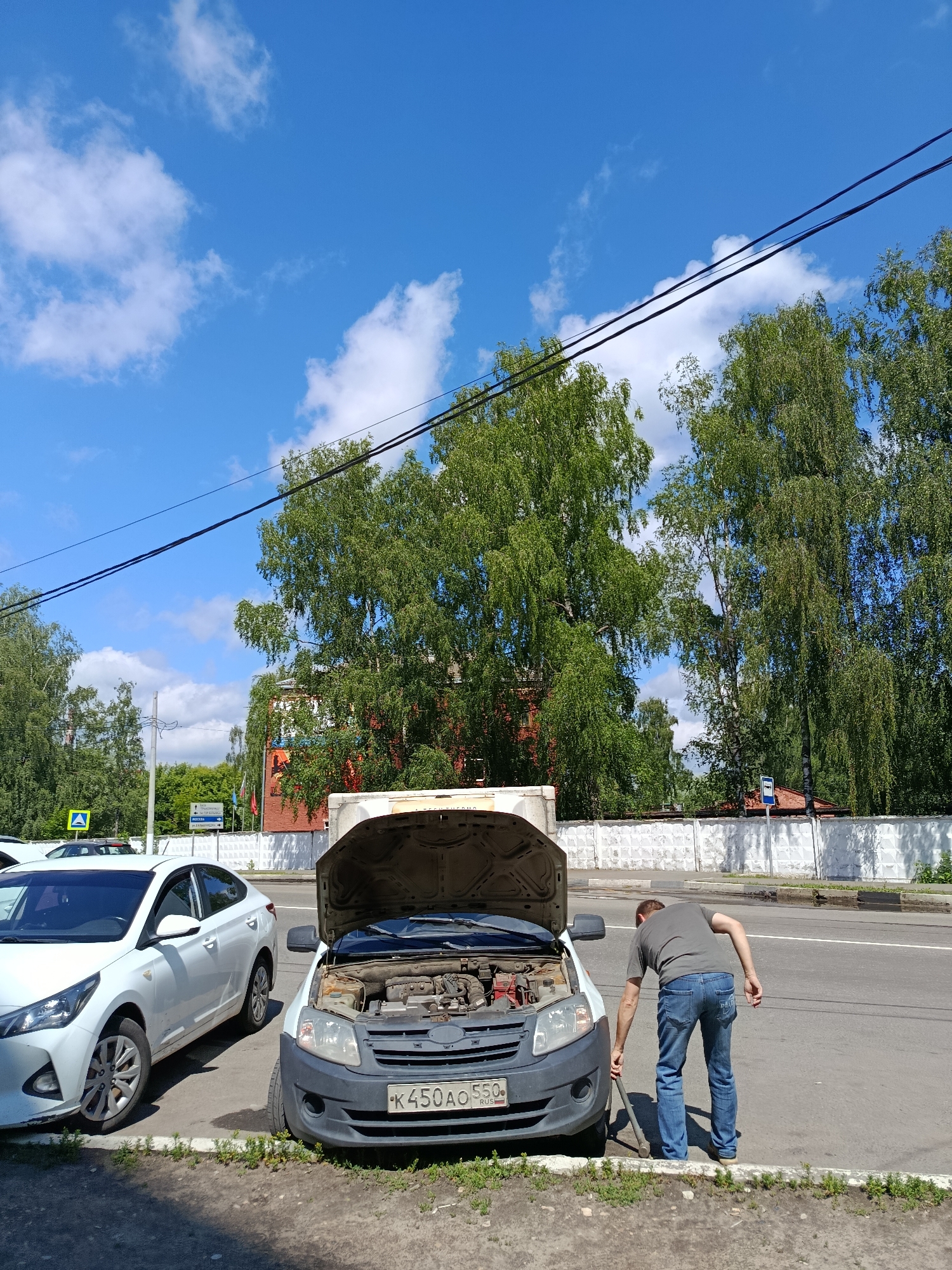 A man in a gray t-shirt and blue jeans is seen from behind, standing on the asphalt next to a car with its hood open. He is using a tire iron to work on the front wheel of the car, which is a light-colored van. Another white sedan is parked to the left of the van. The scene appears to be along a street in Orekhovo-Zuyevo, Russia, with trees and buildings visible in the background under a bright blue sky with scattered clouds. A white fence runs along the side of the street.