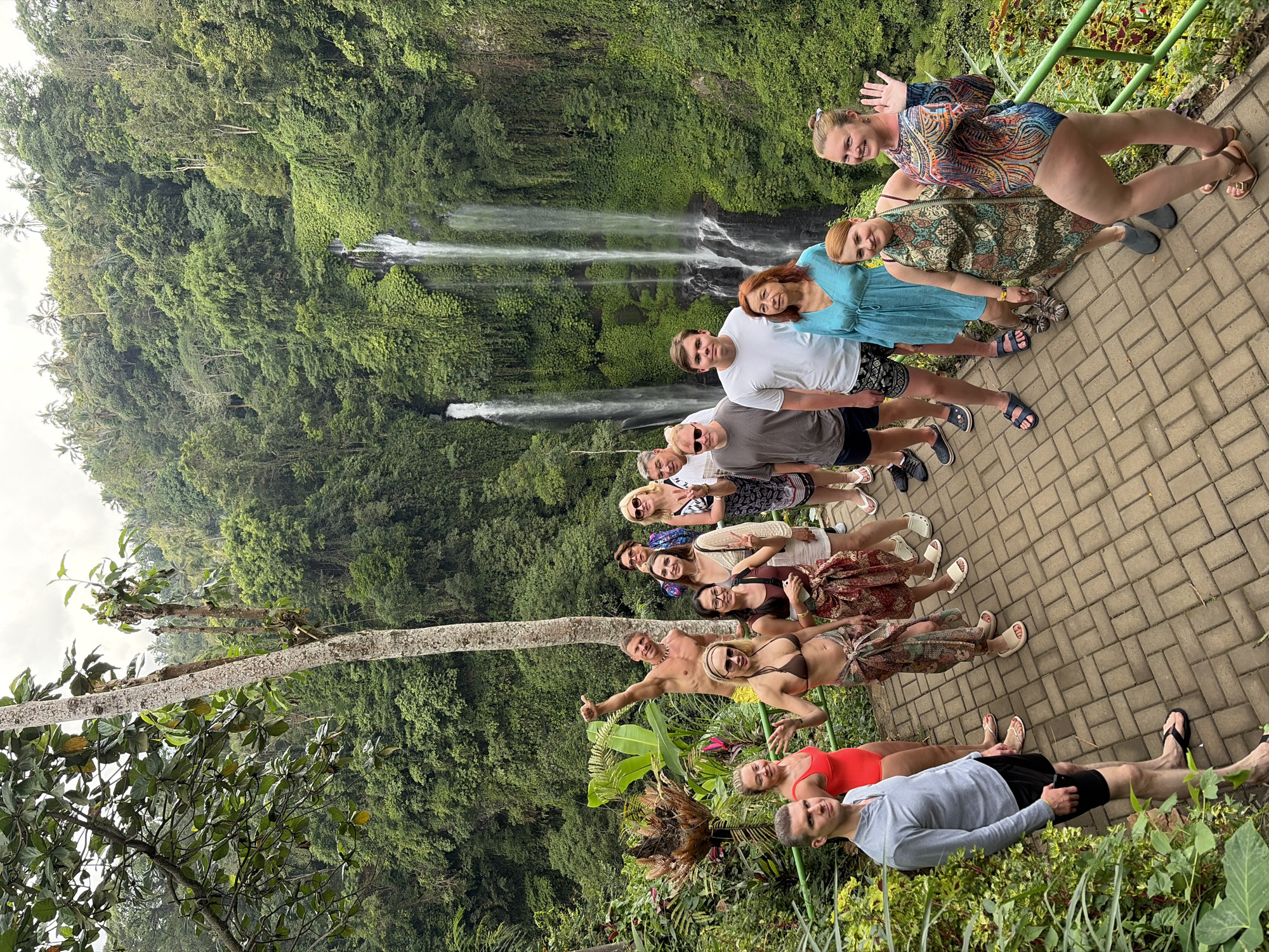 In Sawan, Indonesia, a group of thirteen individuals poses for a photograph on a paved pathway overlooking a lush, green gorge with waterfalls cascading down the cliff face. The group is diverse in age and gender, with most dressed in casual attire, swimsuits, or sarongs suitable for the tropical environment. Some are smiling, waving, or giving a thumbs-up, suggesting a relaxed and enjoyable atmosphere. The background is dominated by dense vegetation, with trees and plants covering the steep slopes. The sky above is overcast. To the right of the pathway, a green metal railing runs along the edge of the gorge.