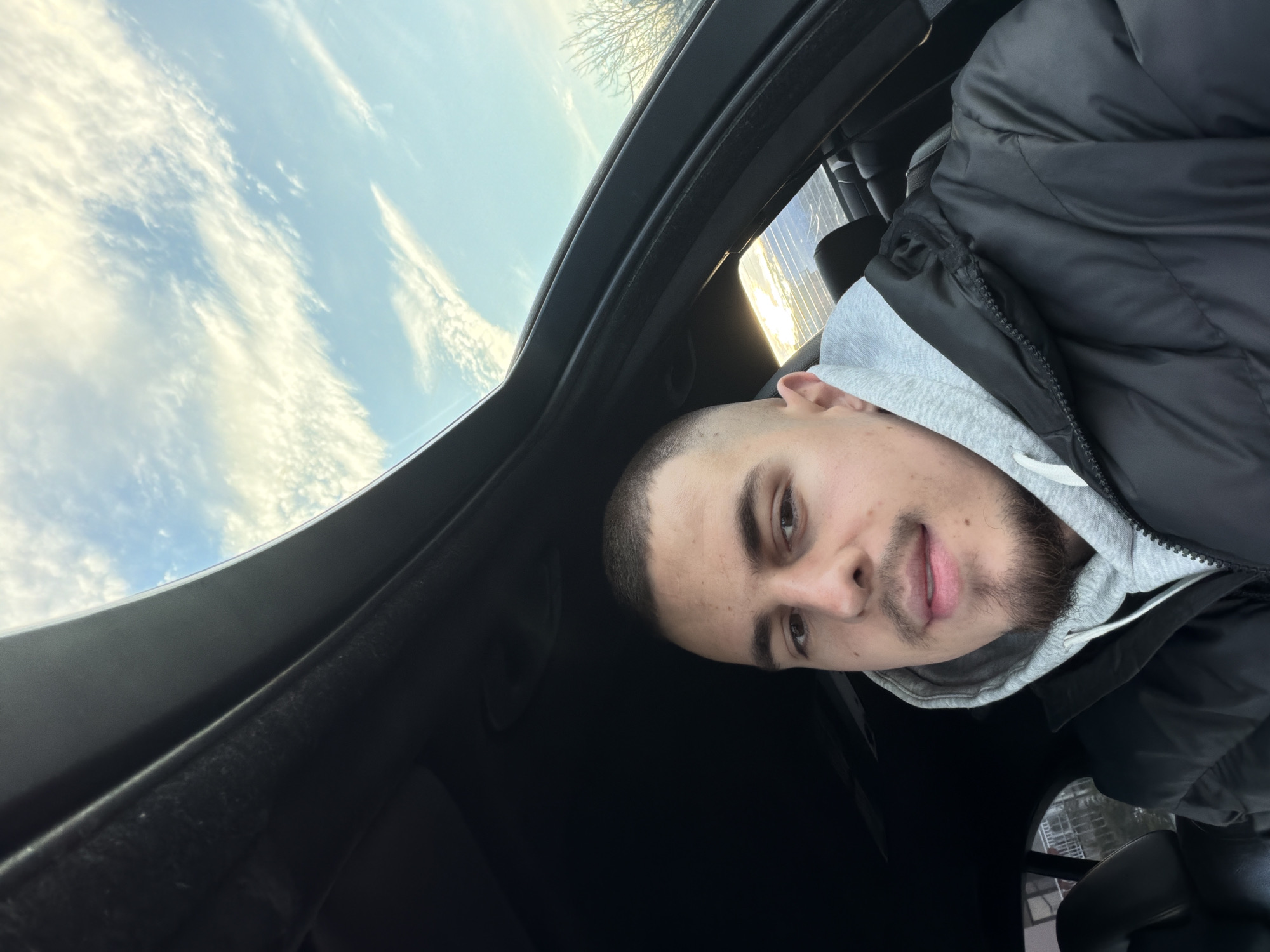 The image shows a man inside a car, possibly looking through the sunroof. The sky is visible through the opening. The man is wearing a grey hoodie and a black jacket. The interior of the car is black. The location cannot be verified as Plovdiv, Bulgaria with the available information. No specific location in Plovdiv can be inferred.