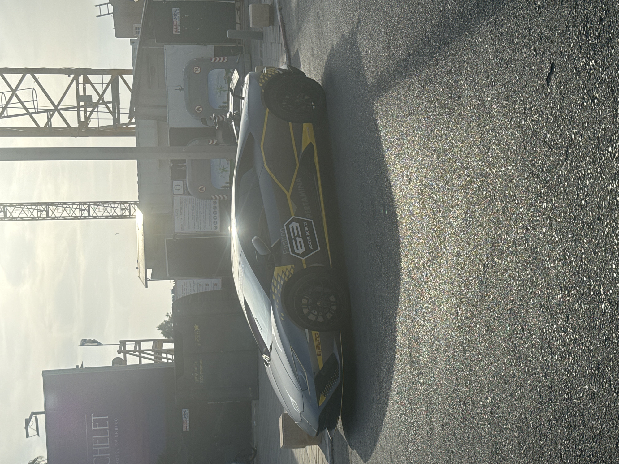 The image depicts a gray Lamborghini Huracan with yellow accents and branding, including "Fastron 63", "ROGER DUBUIS," and "PERTAMINA," parked on a paved surface resembling a street or parking area. The car is oriented towards the viewer's left, casting a shadow onto the street.
In the background, there is construction infrastructure including a crane, and a sign for "CHELET HOTEL BY SHBIRO." Adjacent to this is what appears to be a utility structure with an Israeli flag visible above it. Text on the structure reads "אריזות קרטון משוטחות בלבד הרצל", suggesting the Herzl (Herzliya) area in Israel. The sun is bright in the distance, casting a glare and indicating a daytime scene.