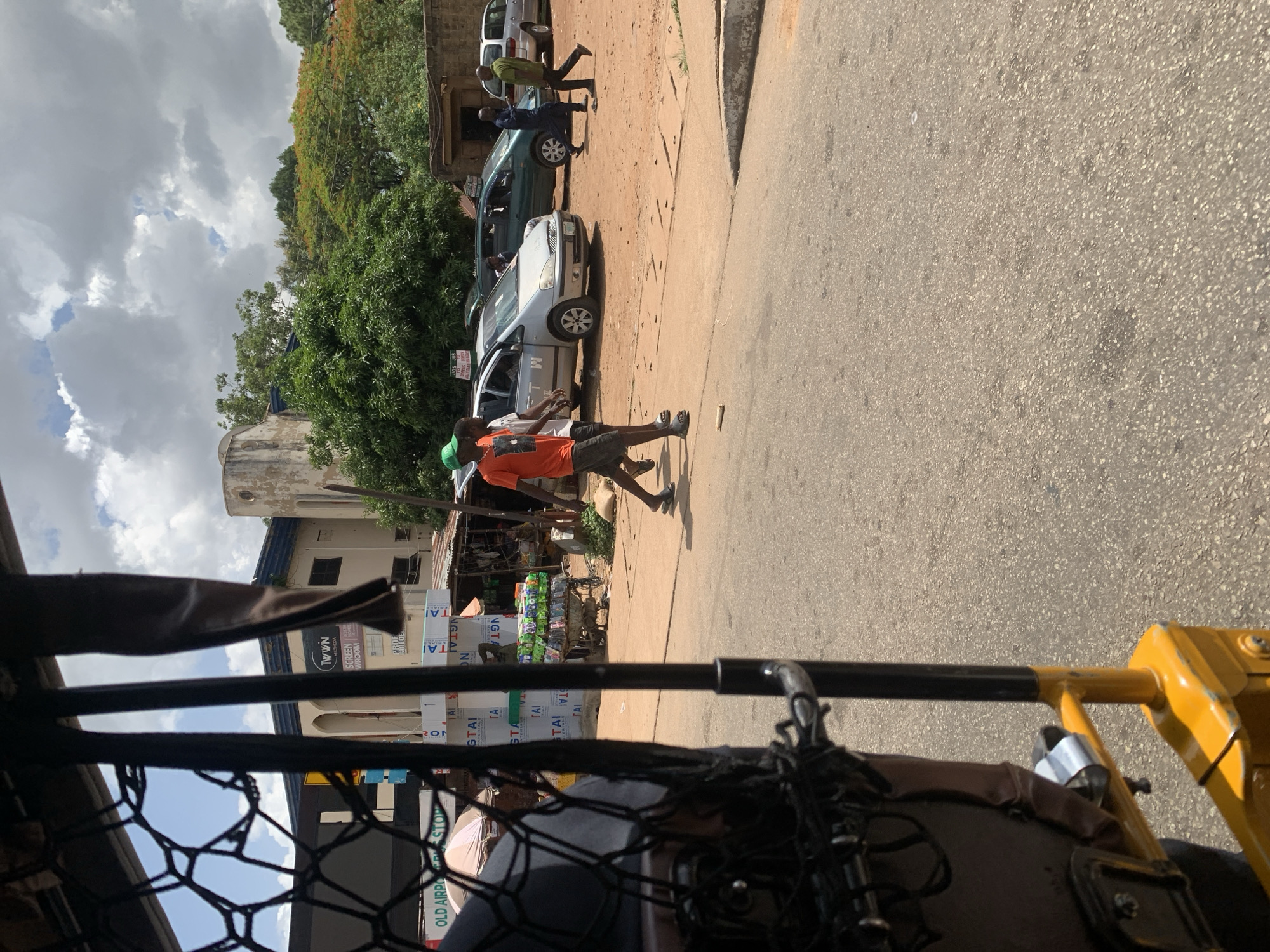 The image presents a vibrant street scene in Jos, Nigeria, captured from inside what appears to be a tricycle or similar vehicle, with its frame and mesh visible in the foreground. The scene depicts a bustling urban or semi-urban environment under a partly cloudy daytime sky.
On the right, an asphalt road runs parallel to a dirt path. Several pedestrians are visible, including a person in an orange shirt and dark shorts walking on the dirt path, and another in a green cap and orange shirt. Farther down the path, an individual in a green shirt stands near a parked dark green sedan. A line of other vehicles, including light grey, silver, and white cars, are parked along the roadside.
On the left side of the street, a vendor's stall is set up, featuring a cart laden with numerous stacked packaged goods, likely drinks or snacks. Large white and green signs prominently display text such as "NGTAI," "GTAI," and "TAI," possibly product branding. Various buildings line the street, one of which features a distinctive rounded structure on its roof. Another building has a sign with "TWWN SCREEN WROOM" (likely "TOWN SCREEN SHOWROOM"). Lush green trees and some with reddish-orange leaves are scattered throughout the background.
Visible text fragments from various signs include "OLD AIRPODS STOP," "ROM," "ON," "PROF," "A," and "1". The bright lighting and active street scene suggest it is daytime.