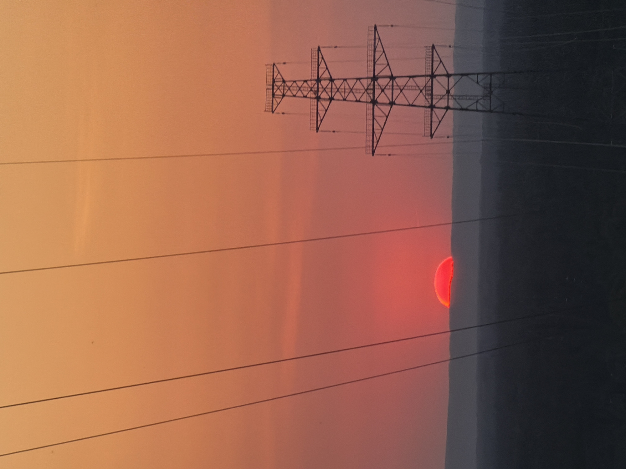 A sideways view of a sunset with a red sun partially obscured by a dark gray landmass. An electrical tower and power lines are silhouetted against the orange and pink sky. The location is likely near Noviy, Russia, given the prompt. The time is likely dusk or dawn due to the sun's position on the horizon.