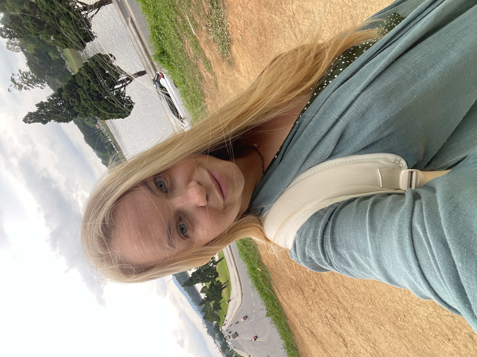 A woman with blonde hair and blue eyes is taking a selfie outdoors in Lâm Viên - Đà Lạt, Vietnam. She is smiling and wearing a teal shawl over a green polka dot dress. She has a light-colored backpack on. The background shows a body of water with trees, a road with traffic, and a cloudy sky. The ground in the foreground is sandy.