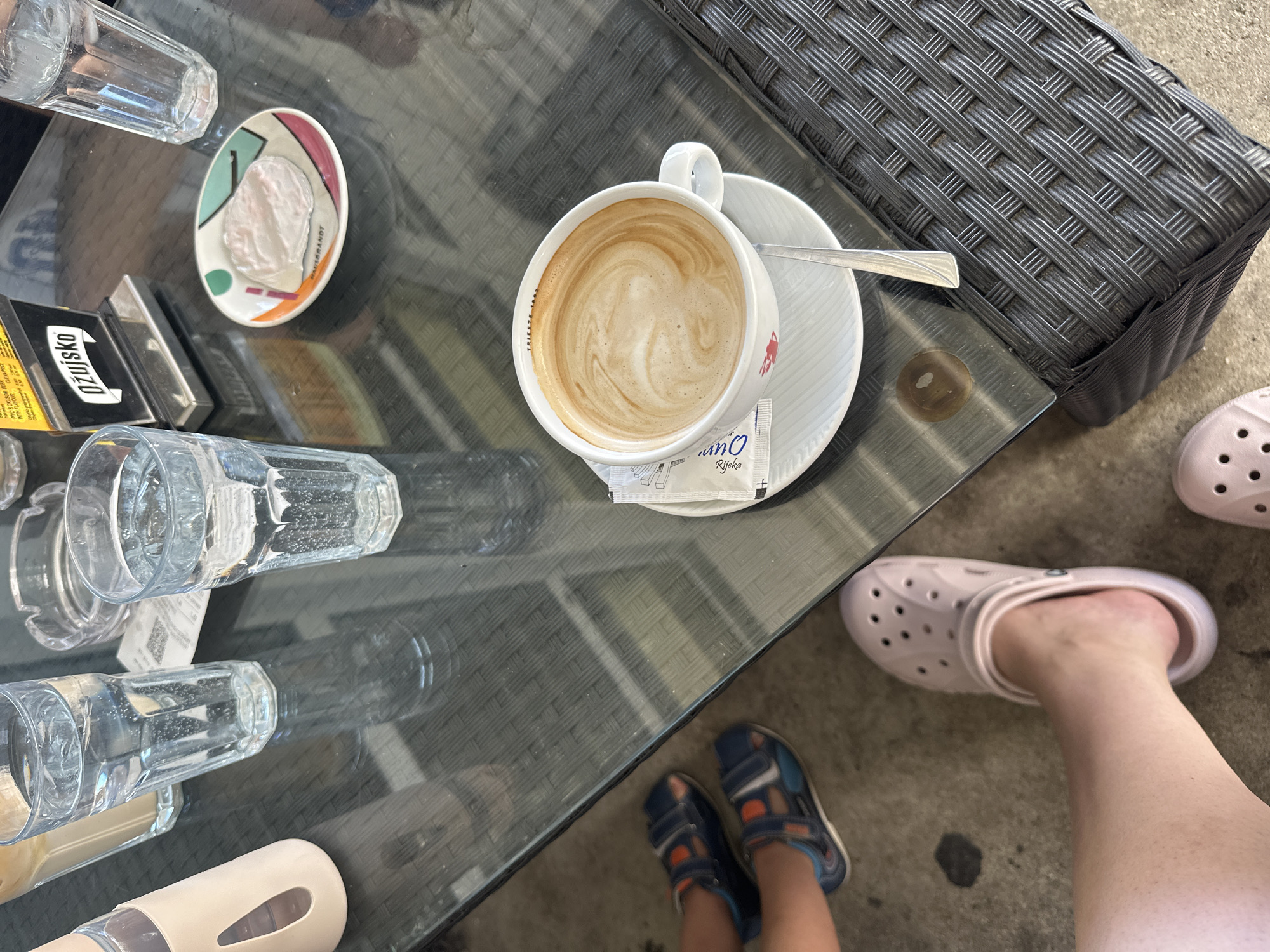 The image depicts a close-up, overhead view of a table at an outdoor cafe in Grad Rijeka, Croatia. The table, made of dark glass with a woven wicker base, holds several items. In the foreground, a white coffee cup with a saucer, containing a cappuccino with latte art, is positioned on the right side. A silver spoon rests on the saucer. To the left of the coffee, a small, decorative plate with a geometric pattern holds a pink confection. Behind the coffee and plate, three glasses of water and an ashtray are visible on the table. On the far left, a light pink reusable water bottle sleeve is partially visible. In the lower left of the image, a person's legs and feet are visible. One foot is wearing a pink Croc sandal, and the other foot is partially obscured by the sandal and a dark blue and orange sandal. The ground beneath the table appears to be concrete, with some dark stains. A woven wicker chair is visible on the right side of the frame. The lighting suggests it is daytime.