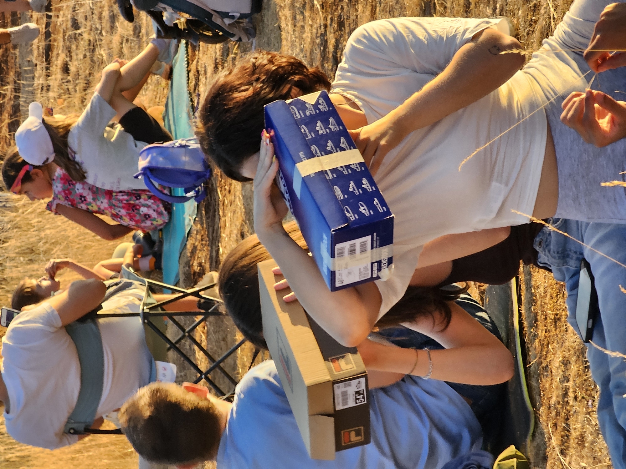 The image depicts a group of people outdoors, likely during daylight with warm, golden lighting suggesting late afternoon or early evening. The setting appears to be a grassy or dry, reedy field, possibly a park or natural area within the city of Schifflange, Luxembourg.
In the foreground, two young individuals are prominent. One, on the left, is holding two shoe boxes, one brown and one blue, up to their face, obscuring it. This person is wearing a blue t-shirt. The other individual, on the right, is also holding a blue shoe box to their face, also obscuring it. This person is wearing a white t-shirt and gray leggings. Their arms are adorned with bracelets and a tattoo.
Behind them, other individuals are visible. To the left, a person is seated in a foldable chair, wearing a white t-shirt. Their back is to the camera. To the right of the chair, a younger person with dark hair is seated. Further back and to the right, two young girls are seated on a blue mat. One wears a colorful floral dress and a pink headband, while the other wears a white t-shirt, black shorts, and a white baseball cap. A blue backpack rests beside them.
The ground is covered in dry grass and some fallen reeds. The overall atmosphere is casual and relaxed, possibly indicating an outing or gathering. Several other objects, including a phone on the ground and parts of what appear to be strollers or outdoor gear, are also present, contributing to the scene of an outdoor recreational activity.
